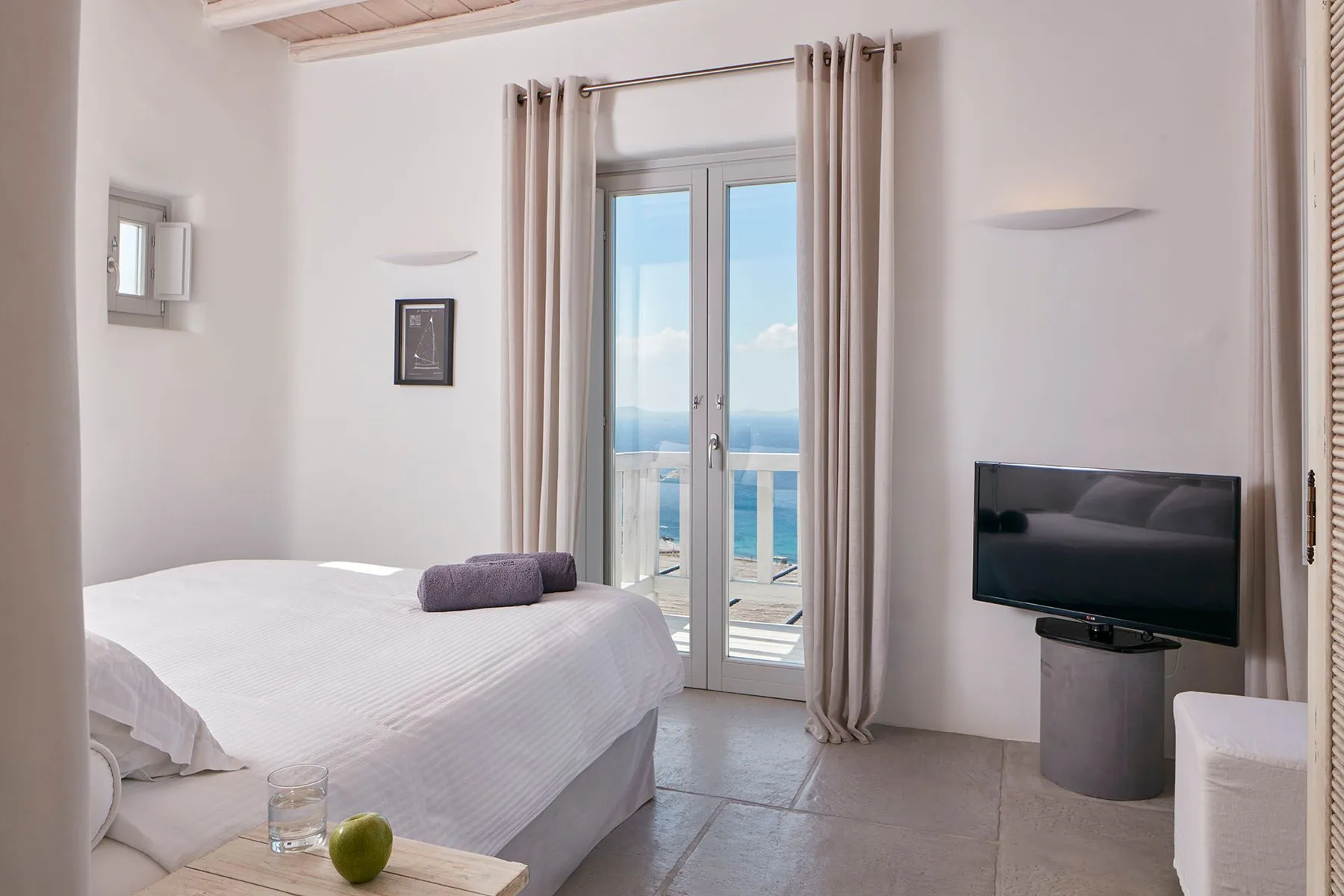 A bright bedroom with a bed, television, and glass doors opening to a sea view.
