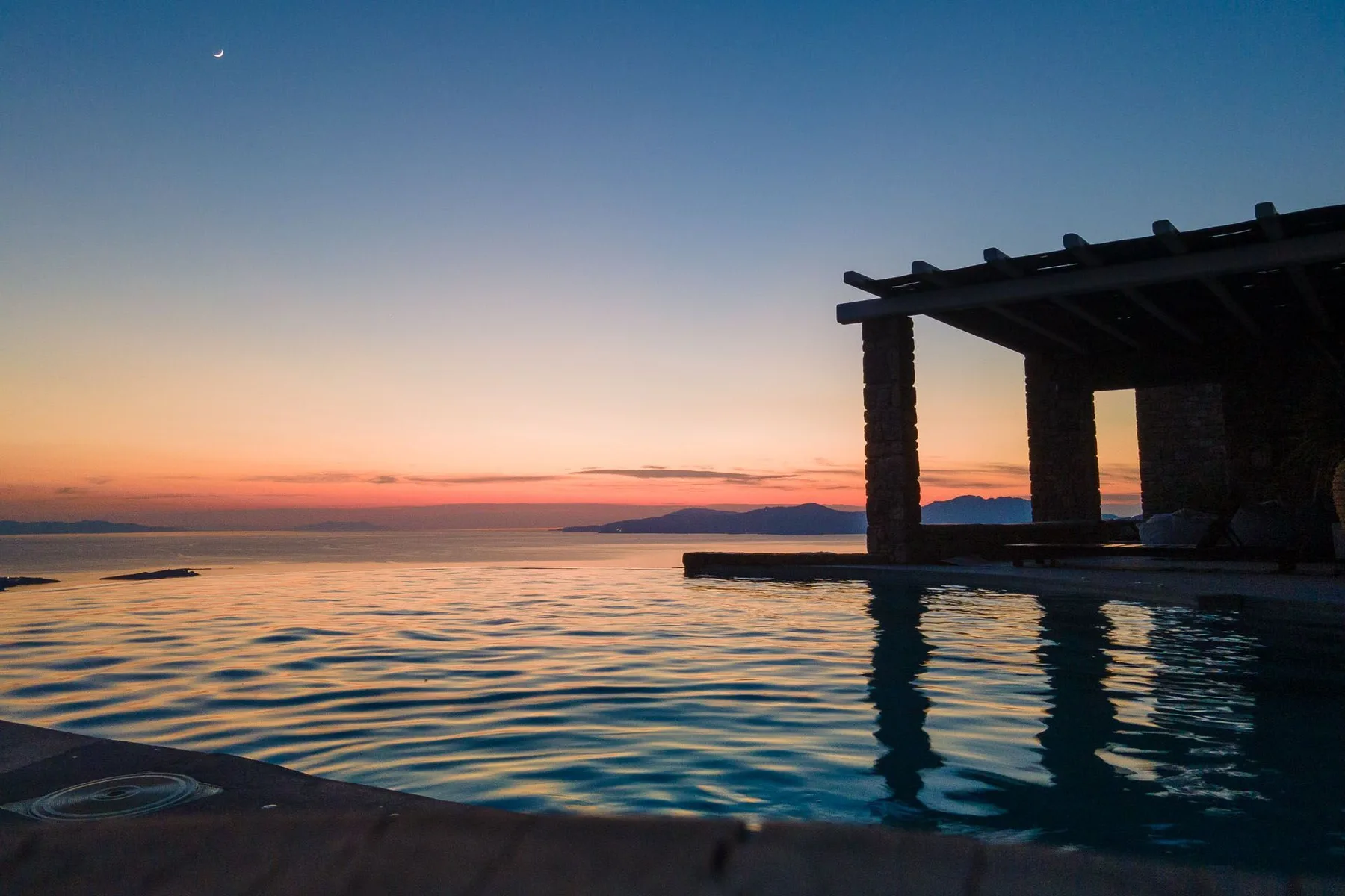 An infinity pool reflects the colors of sunset beside a dark pergola.