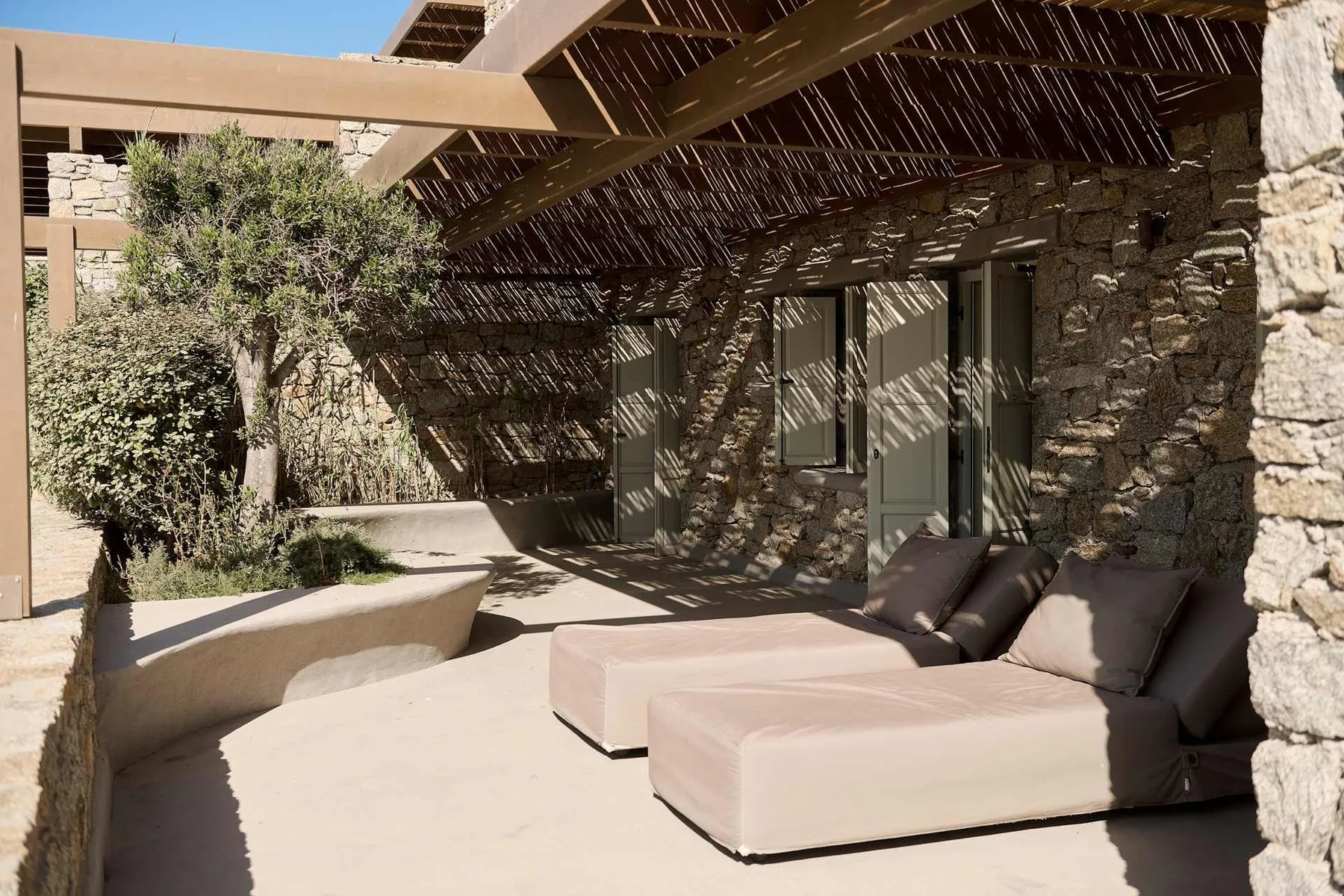 A shaded outdoor seating area with cushioned lounge seating beside a stone wall.