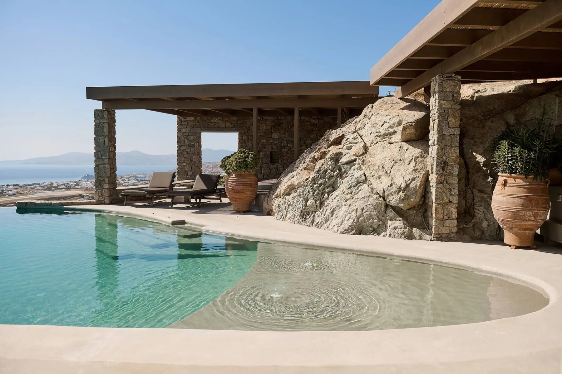 A swimming pool beside a covered patio with lounge seating and rocky landscaping.