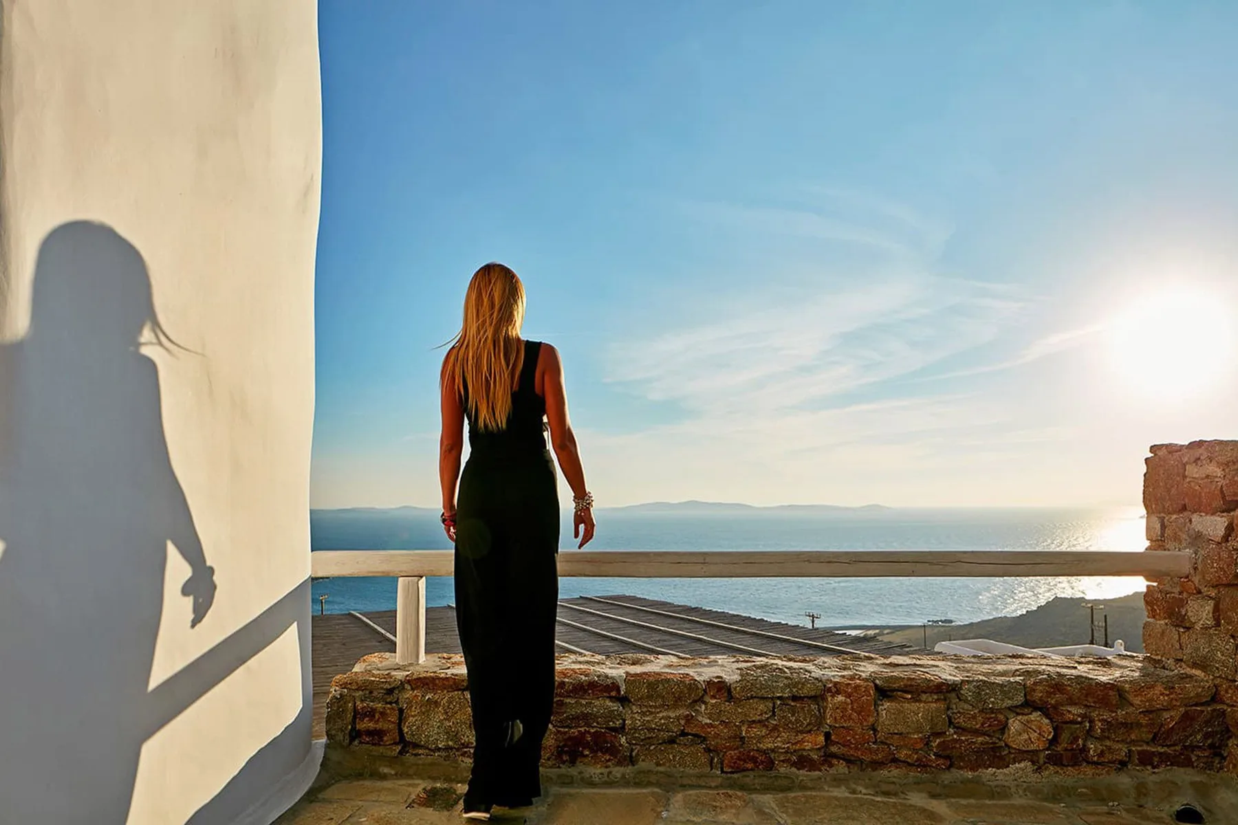A person stands on a stone terrace overlooking the sea in bright sunlight.
