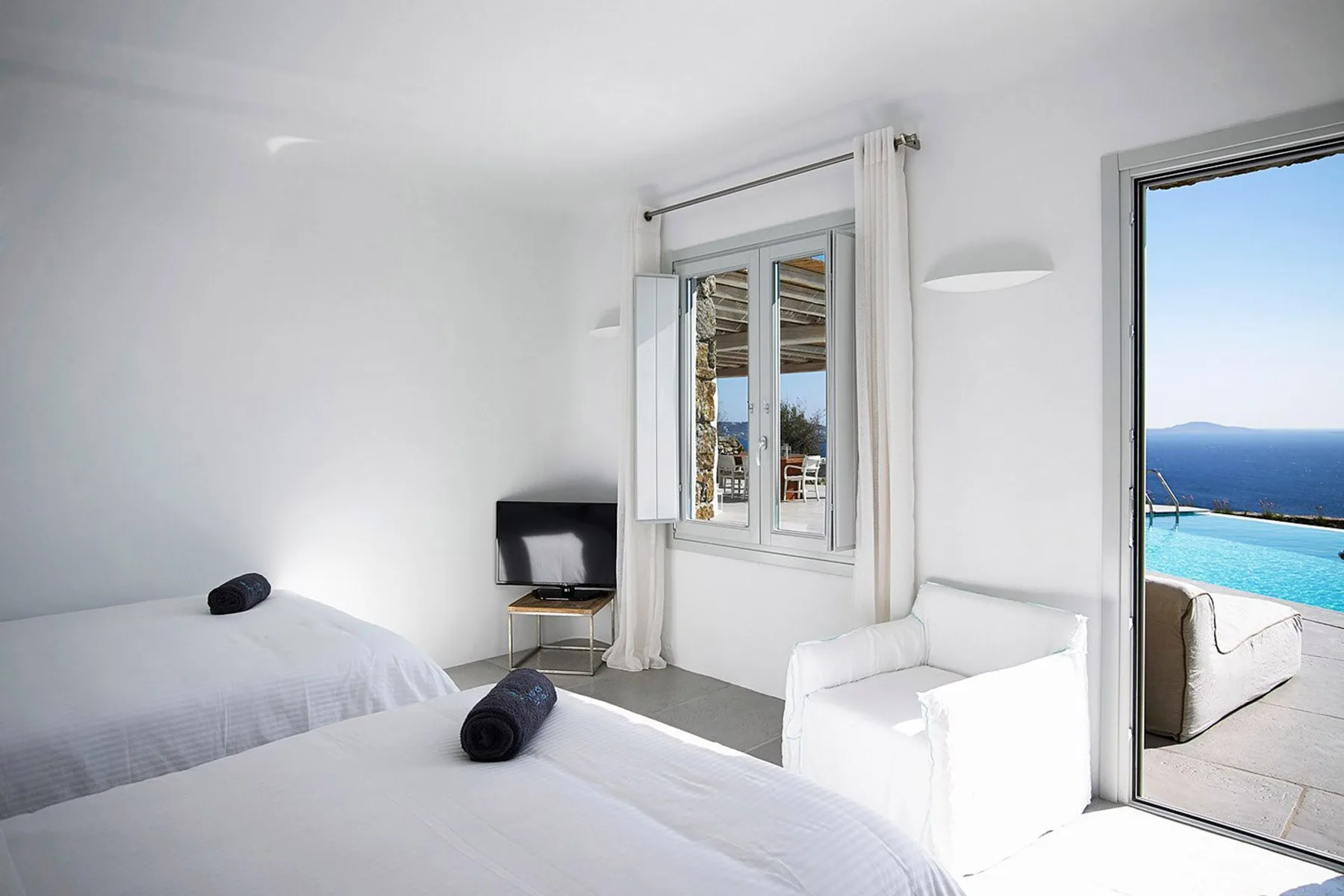A bright bedroom with two beds, a chair, and a view of the sea.