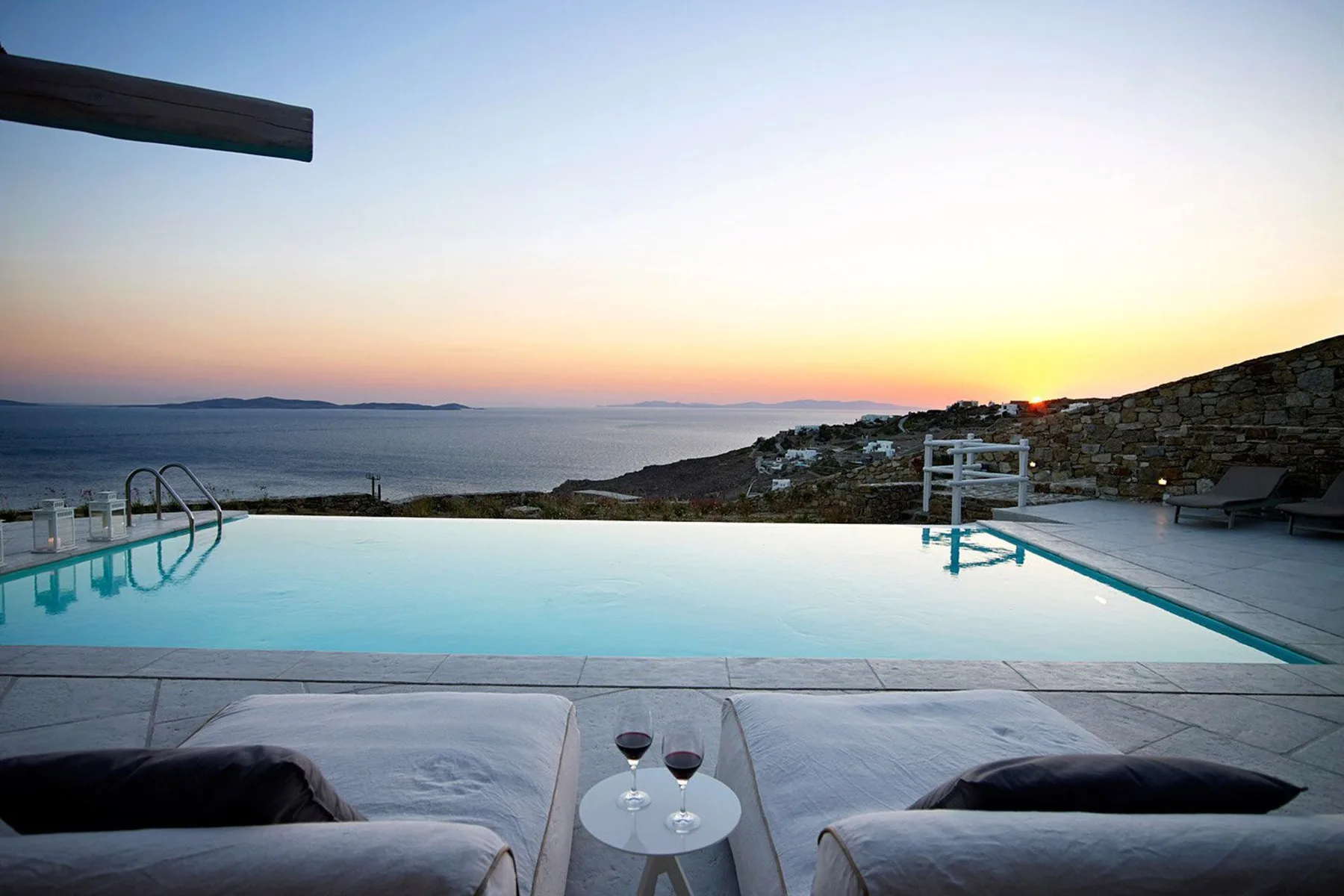 An infinity pool overlooks the sea at sunset, with lounge seating in the foreground.