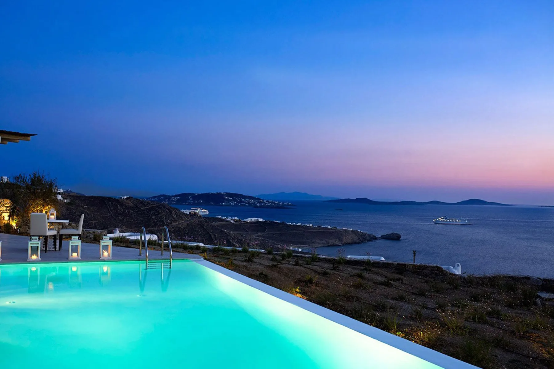 An illuminated swimming pool overlooks the sea at dusk.