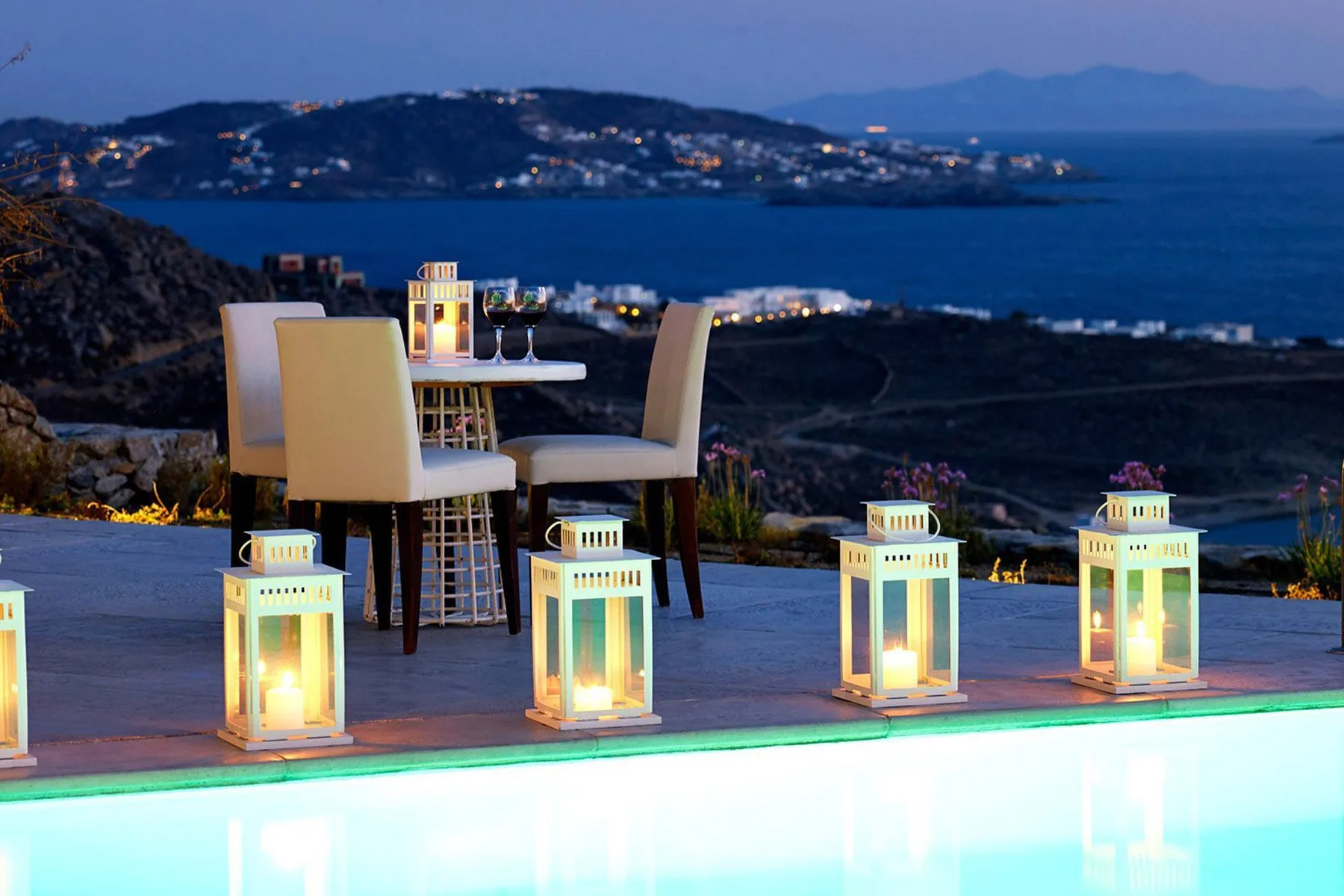 An outdoor dining area beside a lit pool overlooks the sea at dusk.