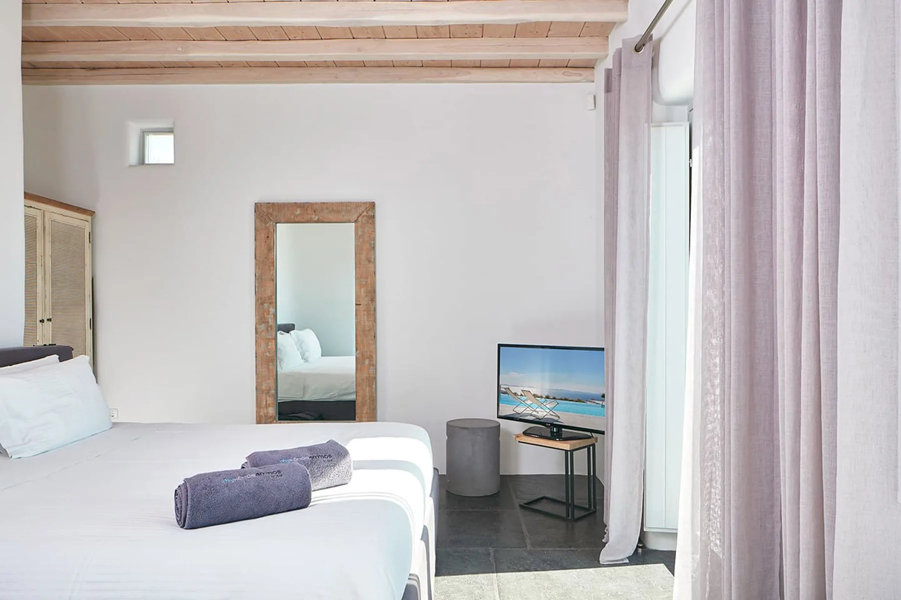 A bedroom with a bed, a full-length mirror, and curtains beside a bright window.