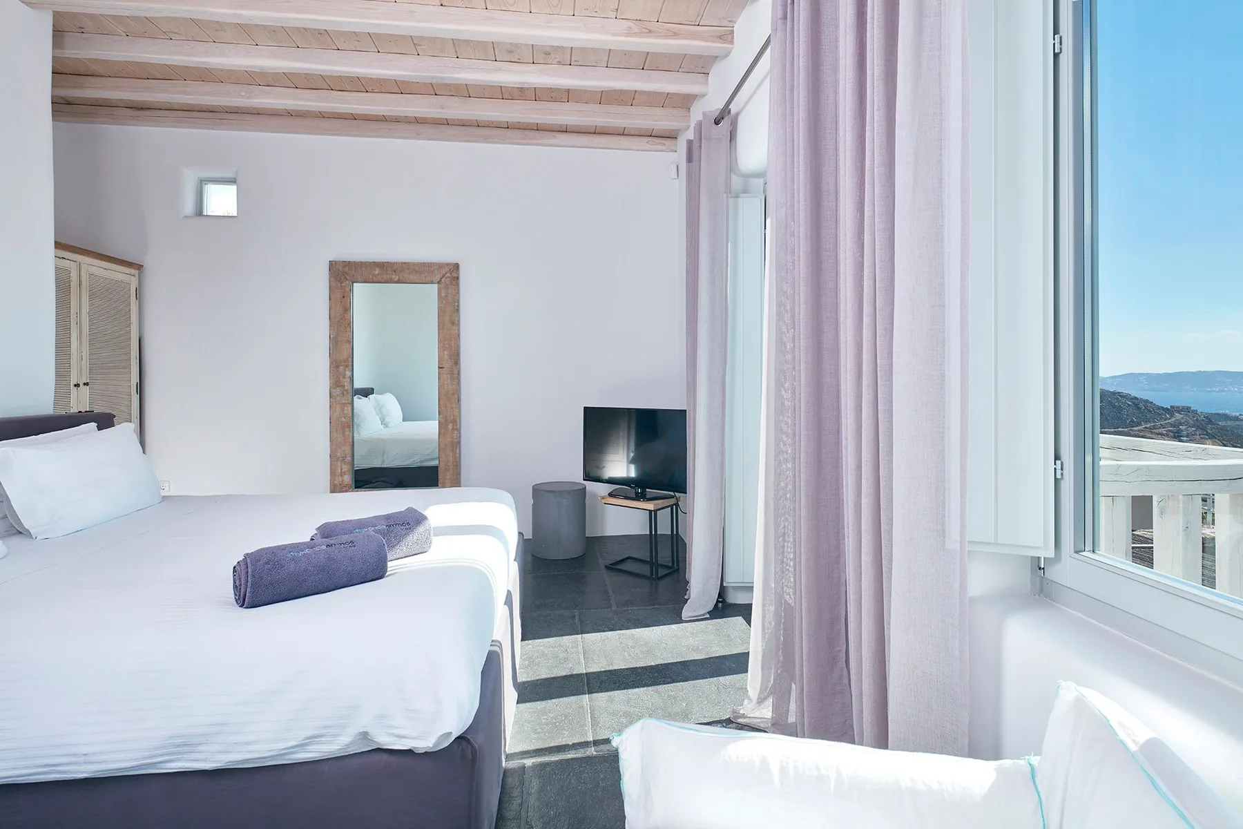 A bright bedroom with a bed, television, and windows overlooking the coast.