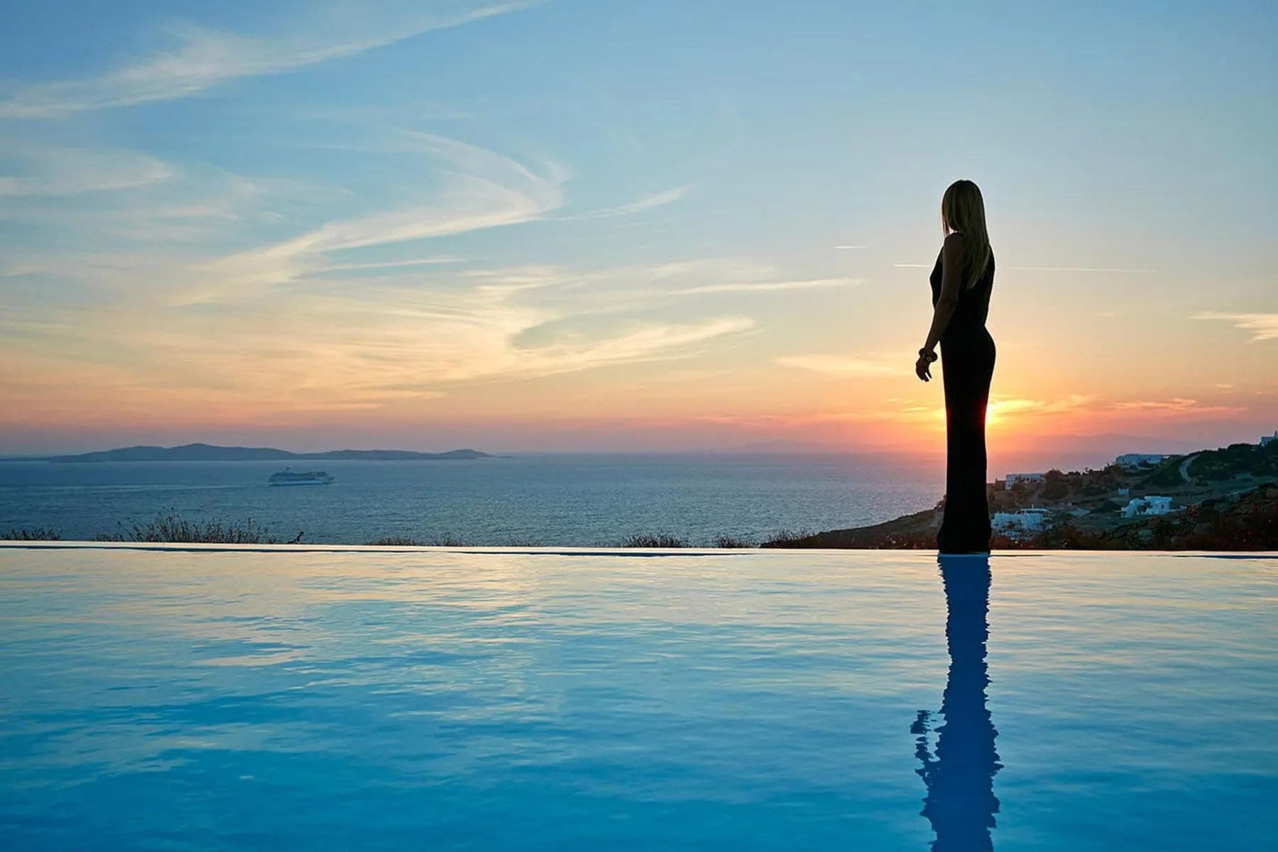 A person stands beside an infinity pool overlooking the sea at sunset.
