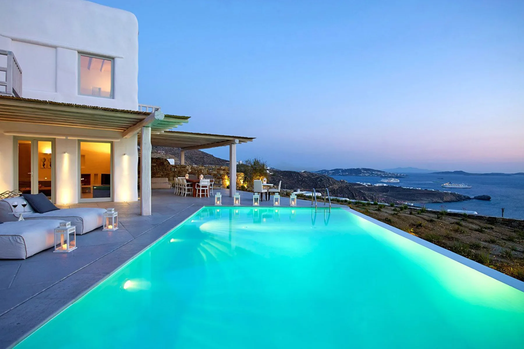 A modern house overlooks a lit swimming pool and the sea at dusk.