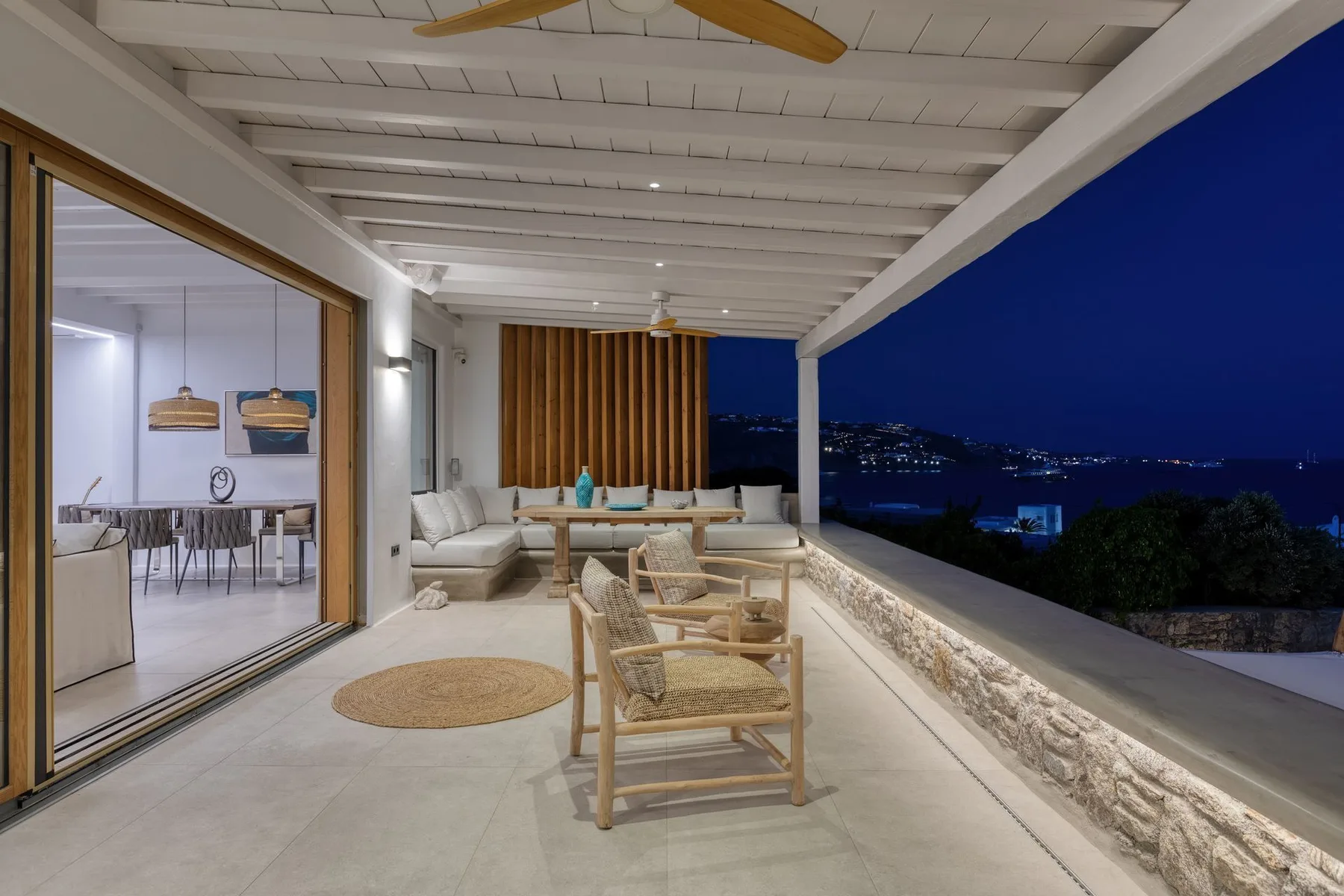 A covered terrace with wicker seating overlooks a distant city at night.