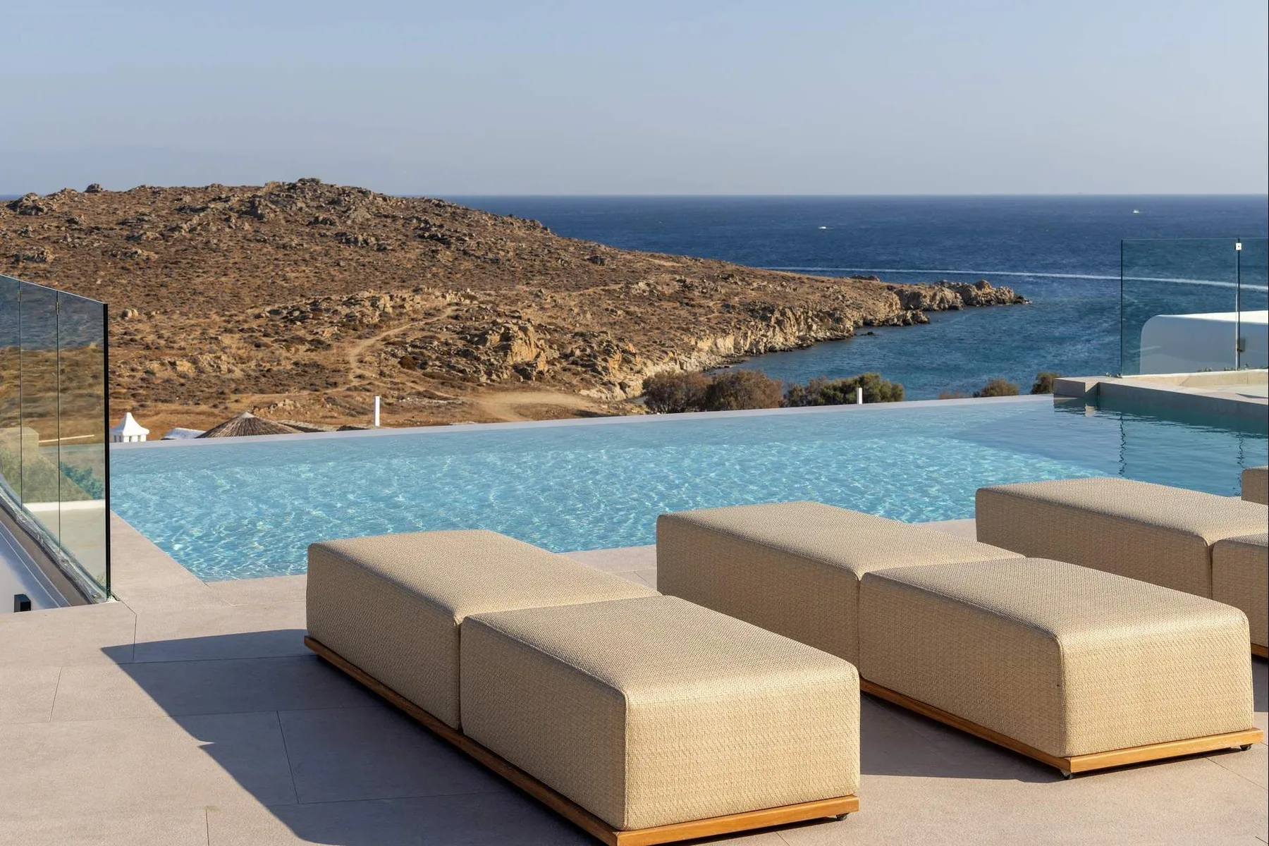 An outdoor swimming pool overlooks a rocky coastline, with cushioned seating nearby.