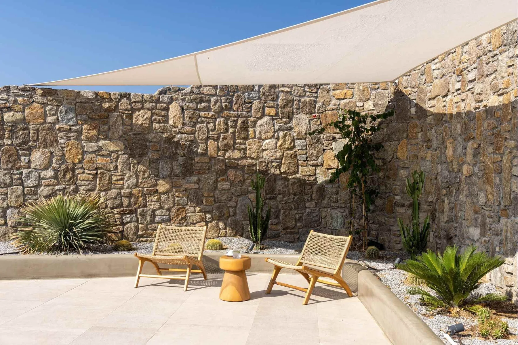 A sunny stone-walled patio with seating beneath a fabric shade.