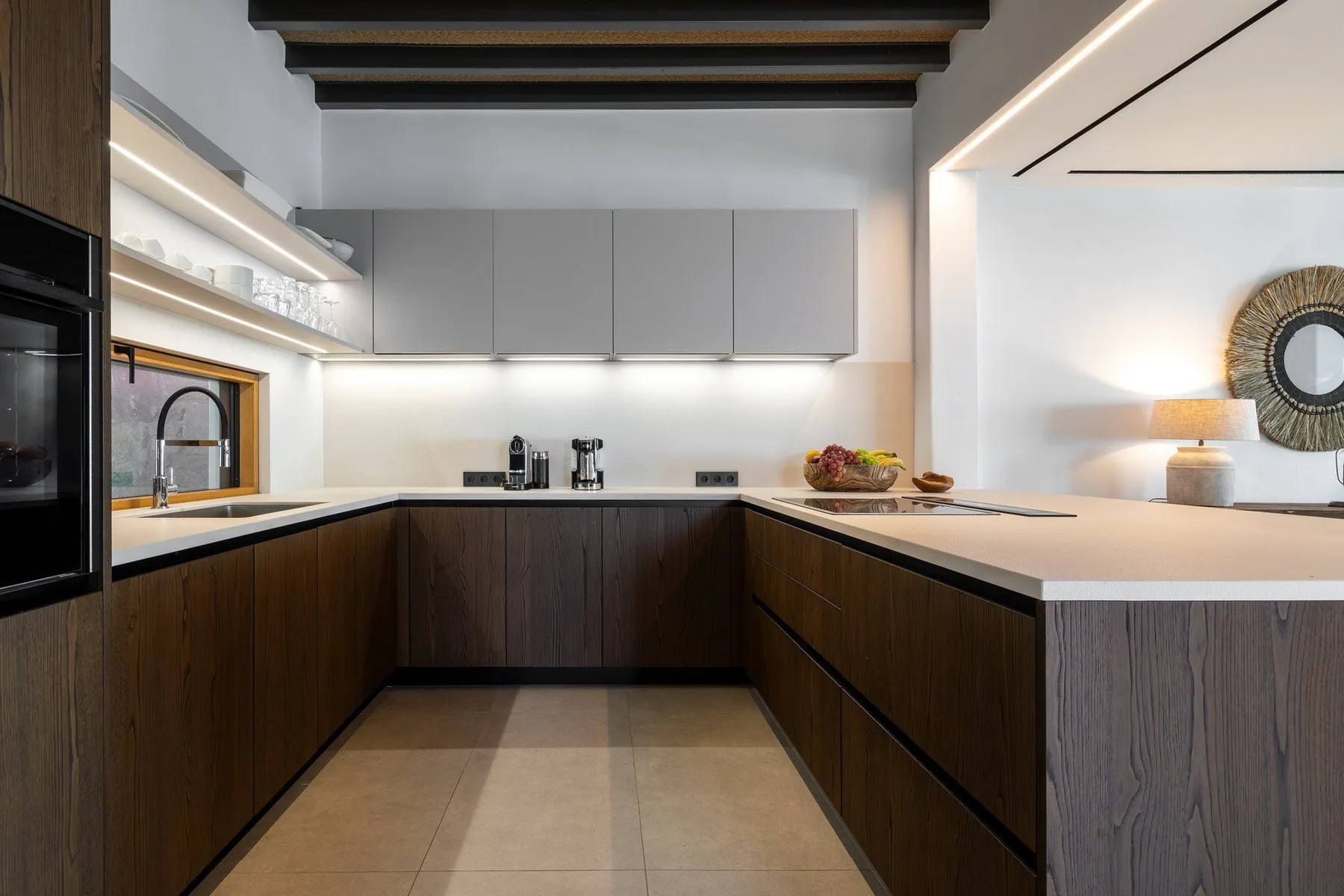 A modern kitchen with dark cabinetry, white counters, and a small sink window.