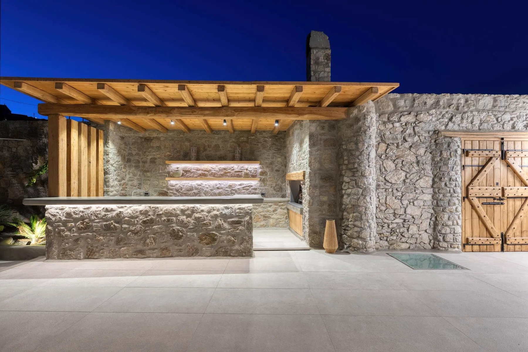 A stone outdoor kitchen beneath a wooden canopy, with a broad counter and paved patio.