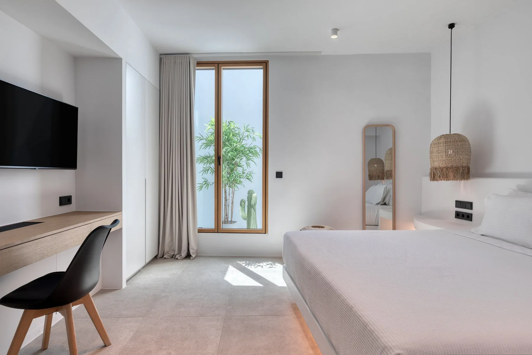 A bright bedroom with a large bed, wall-mounted television, and tall window.