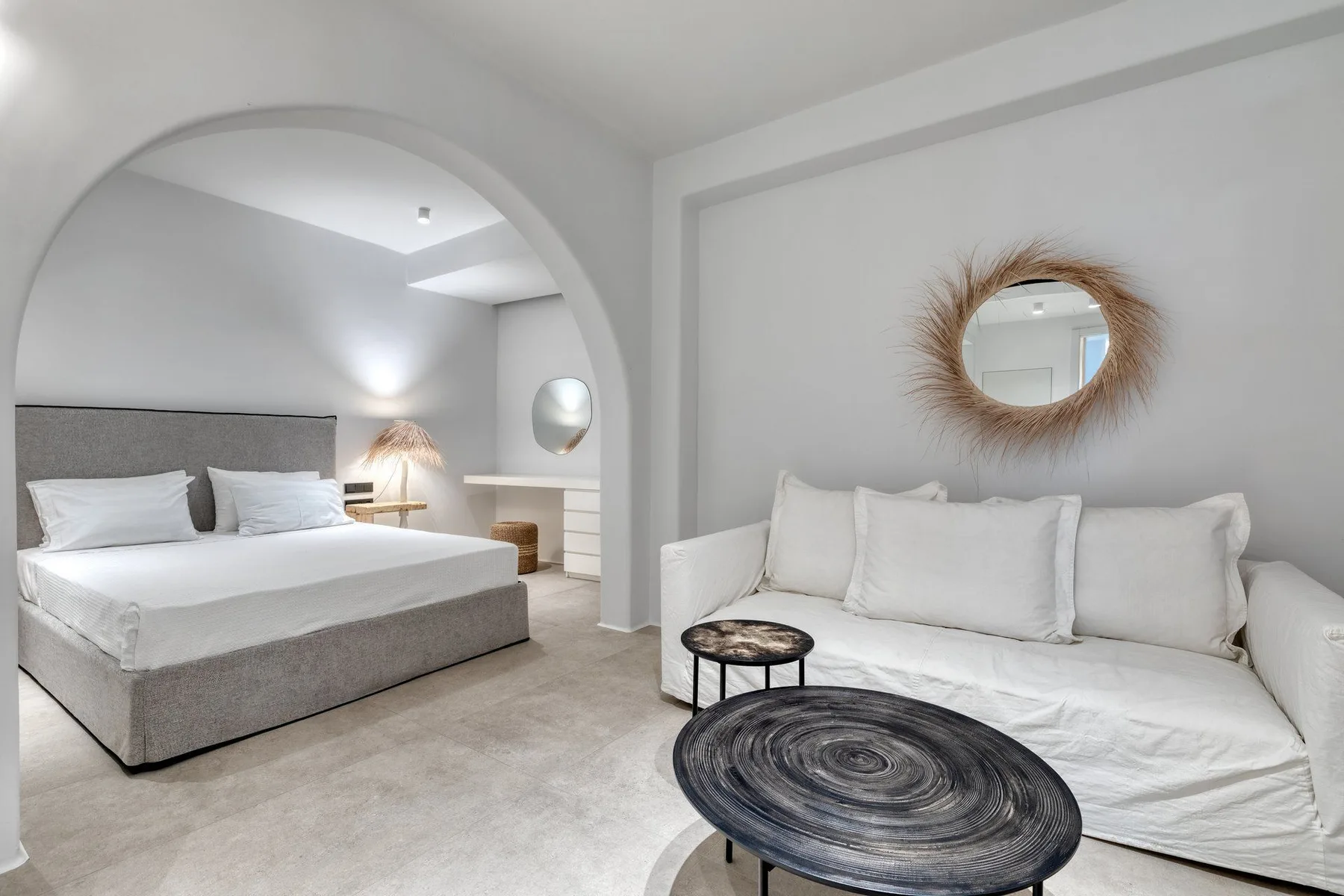 A bright bedroom with a bed and a white seating area beneath a round mirror.