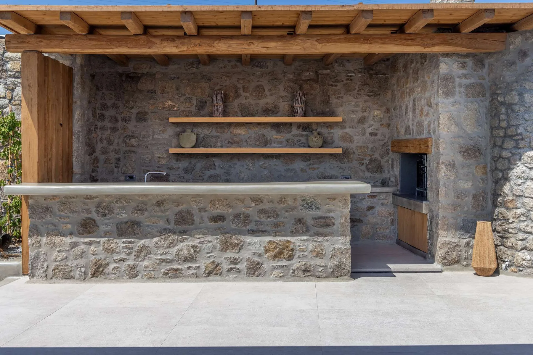 An outdoor stone bar counter sits beneath a wooden pergola.