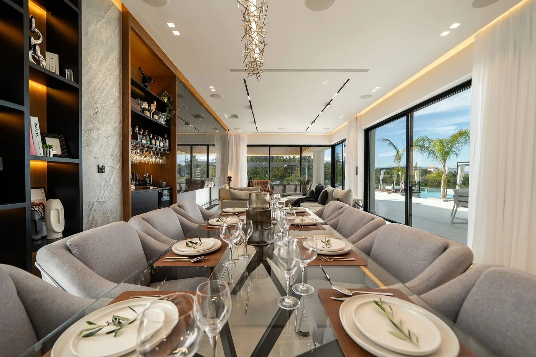 A long dining table is set with plates and glassware beside a lounge area and floor-to-ceiling windows.
