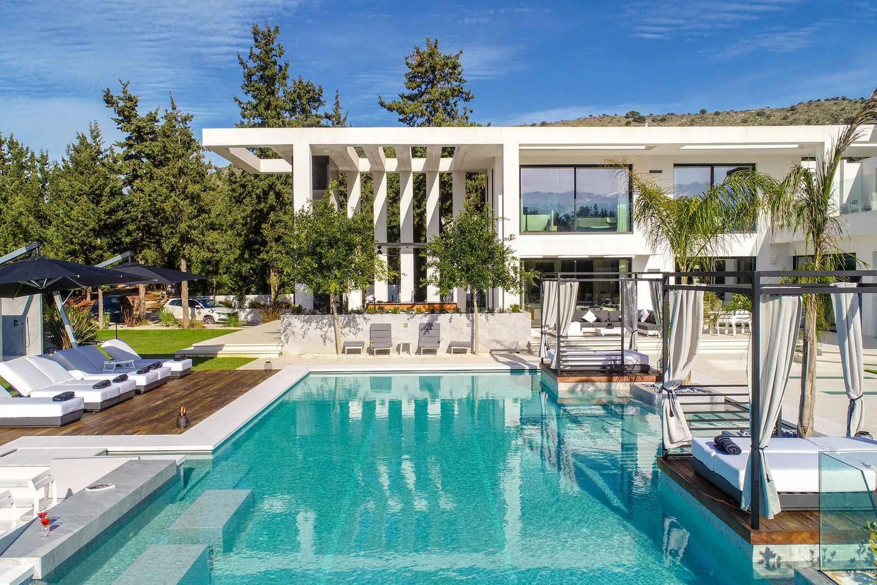 Modern white villa with large windows overlooking a turquoise swimming pool and furnished sun terrace.