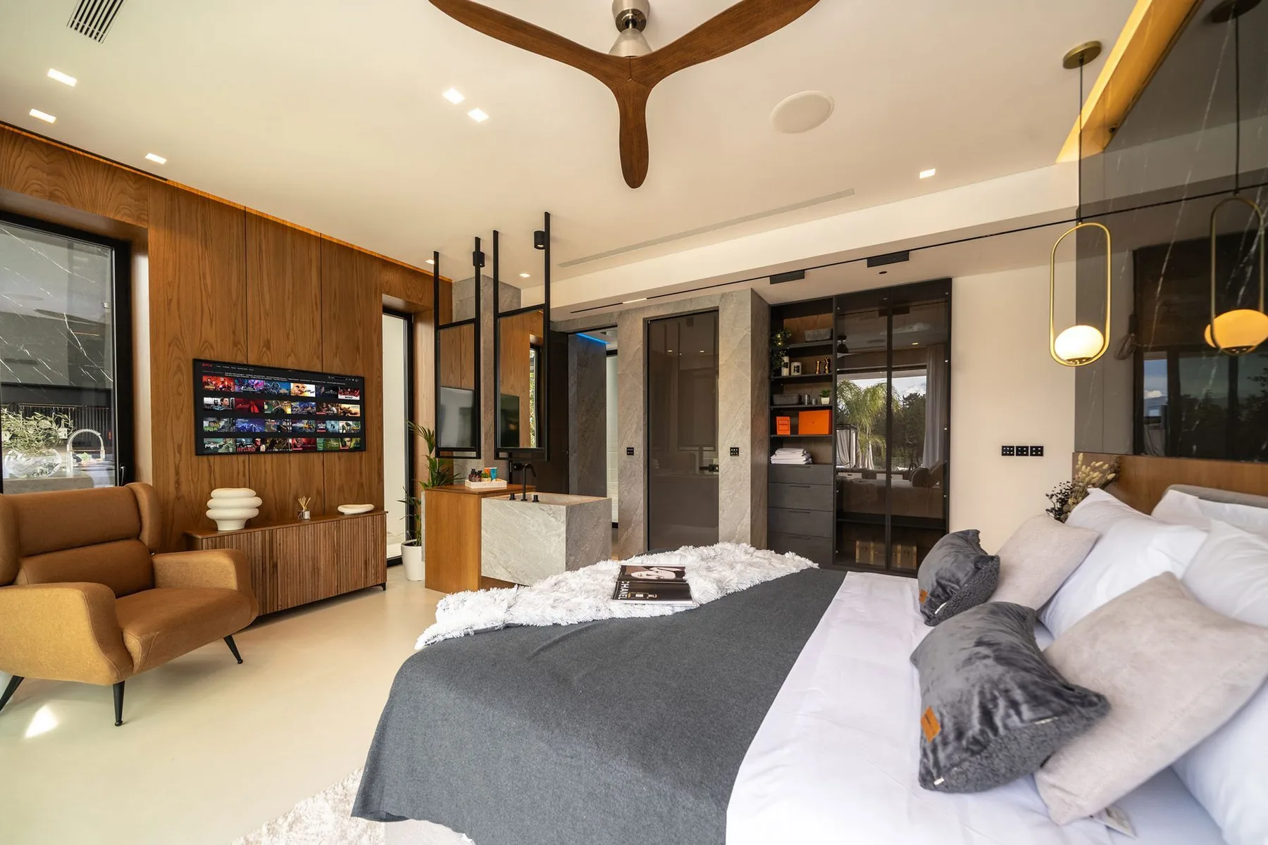 A modern bedroom features a gray bed, tan lounge chairs, wood paneling, and built-in shelving.