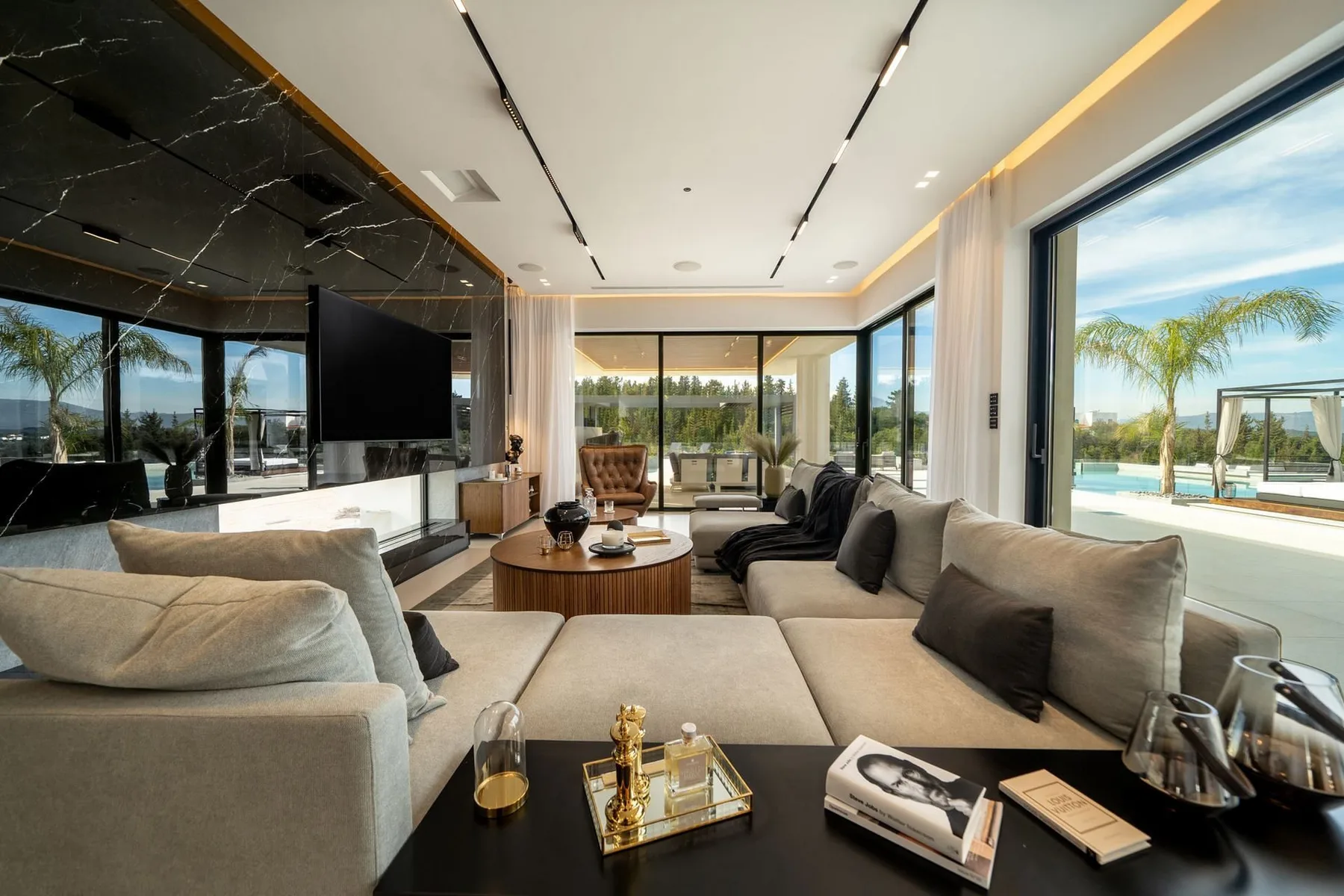 A modern living room features large sectional sofas, a round coffee table, and floor-to-ceiling windows.