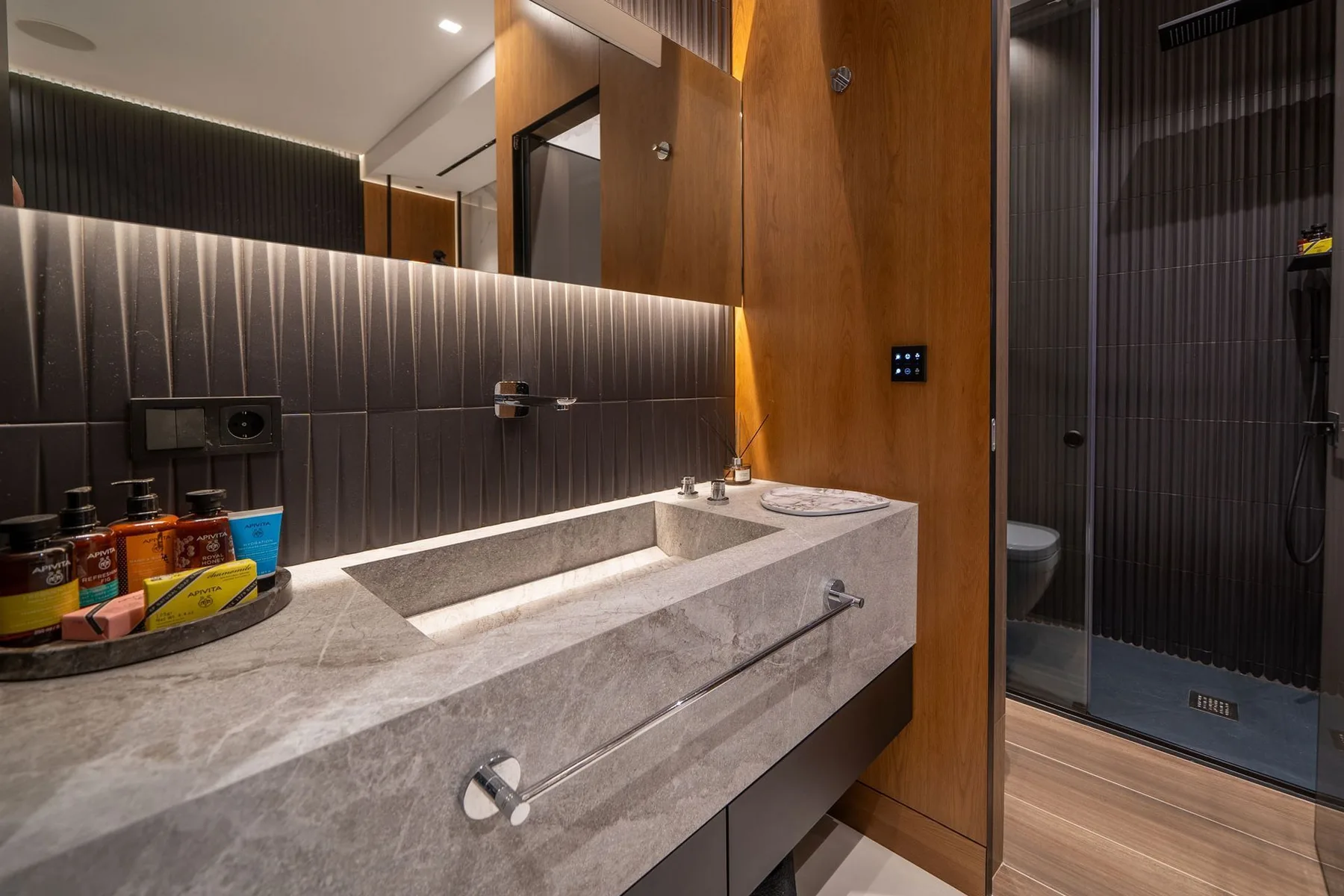A modern bathroom has a long stone vanity, rectangular basin, and dark tiled shower.