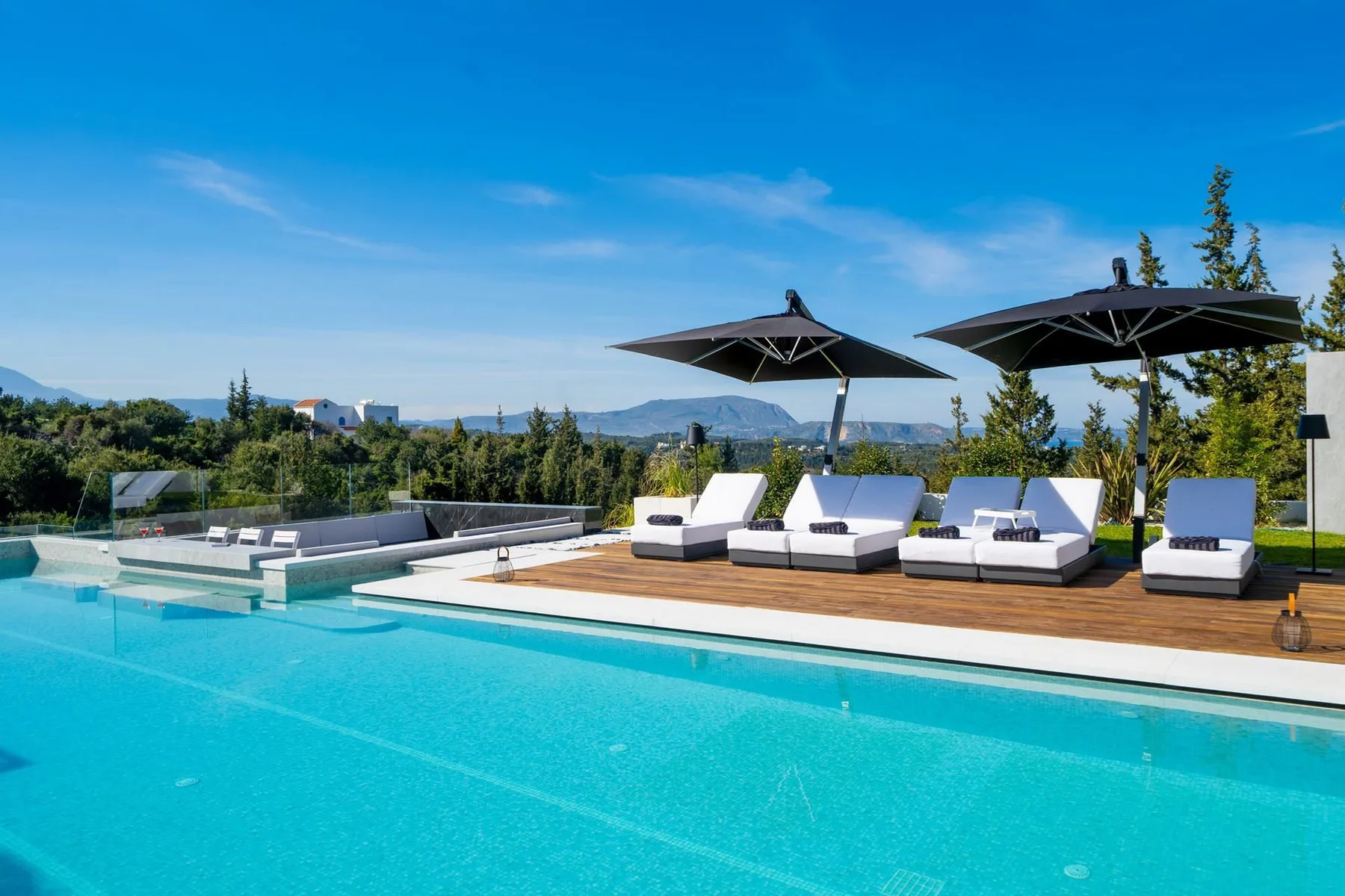 An outdoor swimming pool has white loungers and black umbrellas on a wooden deck.