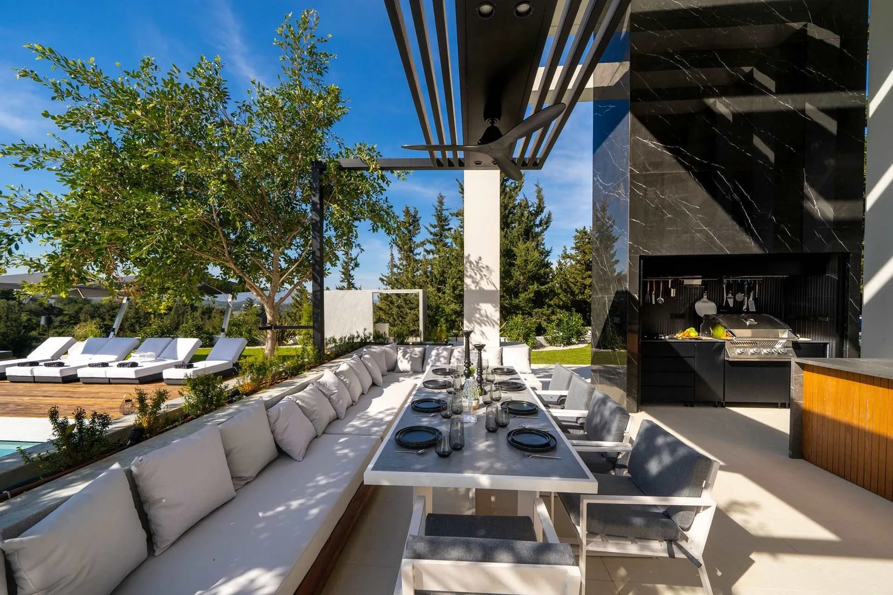 An outdoor dining area features a long table, cushioned seating, a pergola, and a built-in grill.