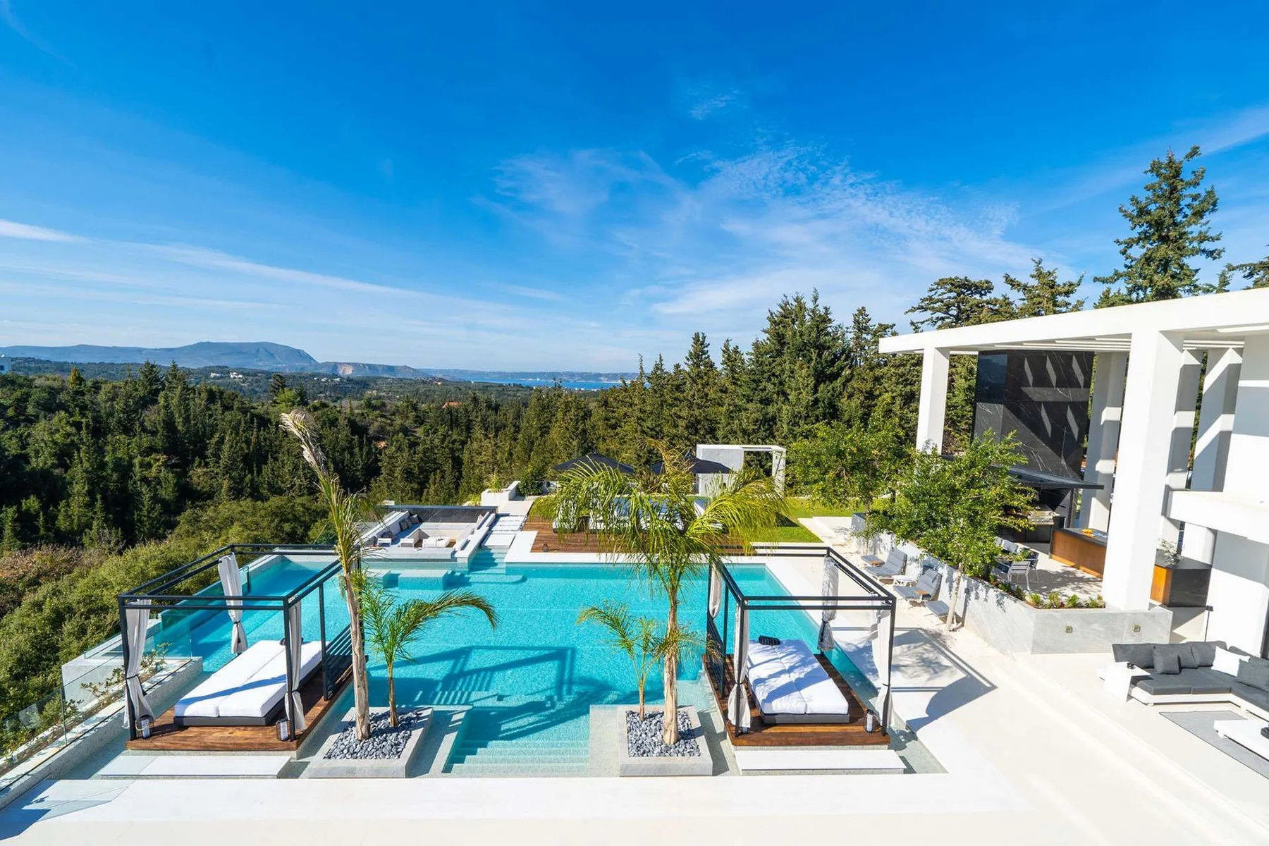 A modern outdoor pool with glass railings sits beside a white pavilion, overlooking a forested landscape.
