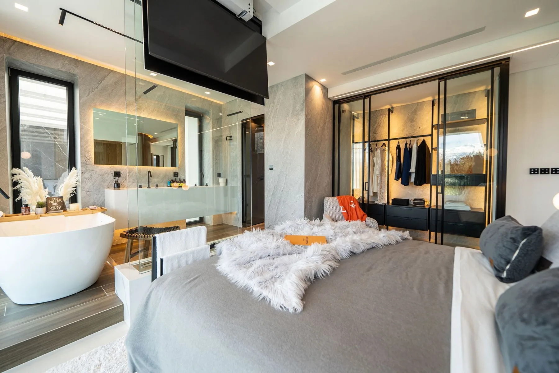 A modern bedroom with a gray bed, freestanding bathtub, glass shower, and illuminated wardrobe.
