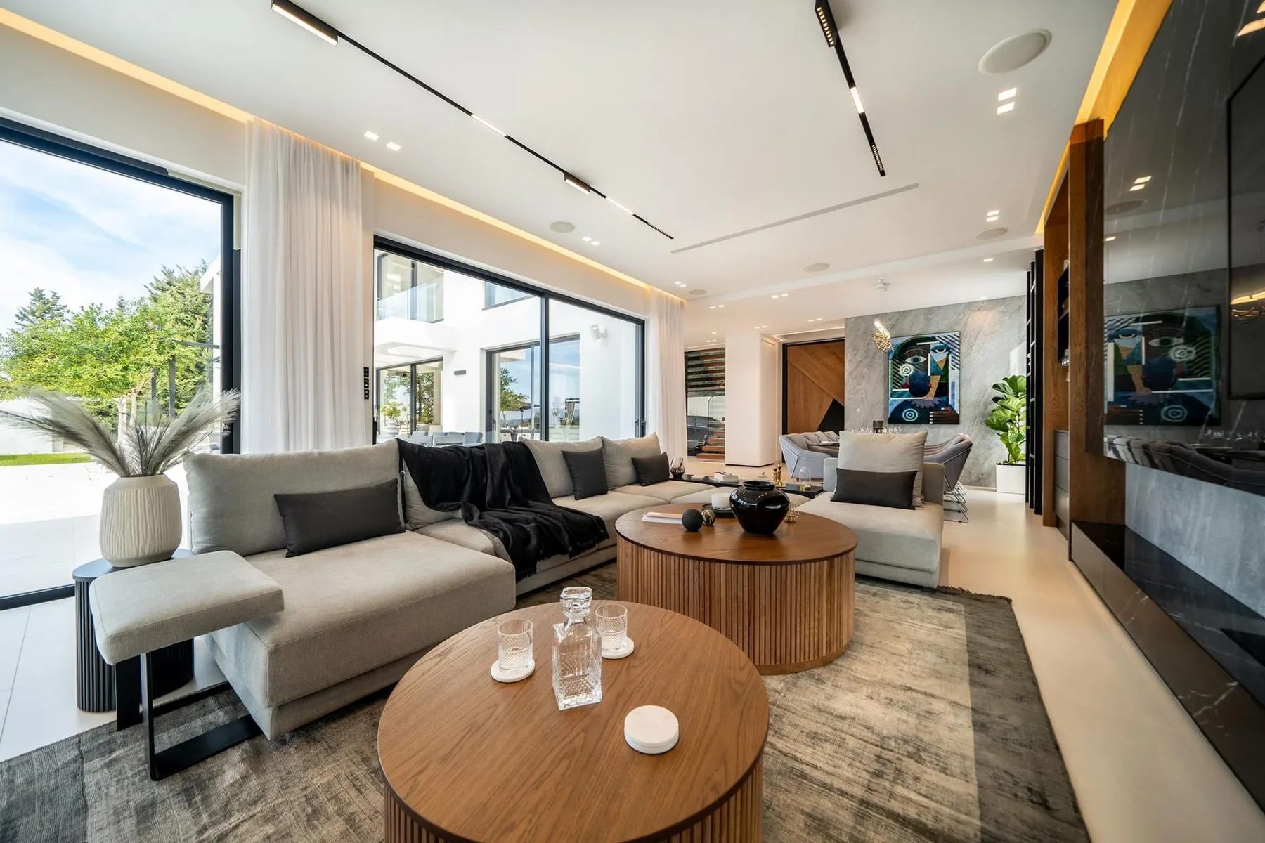 A bright modern living room features large windows, sectional sofas, and round wooden coffee tables.