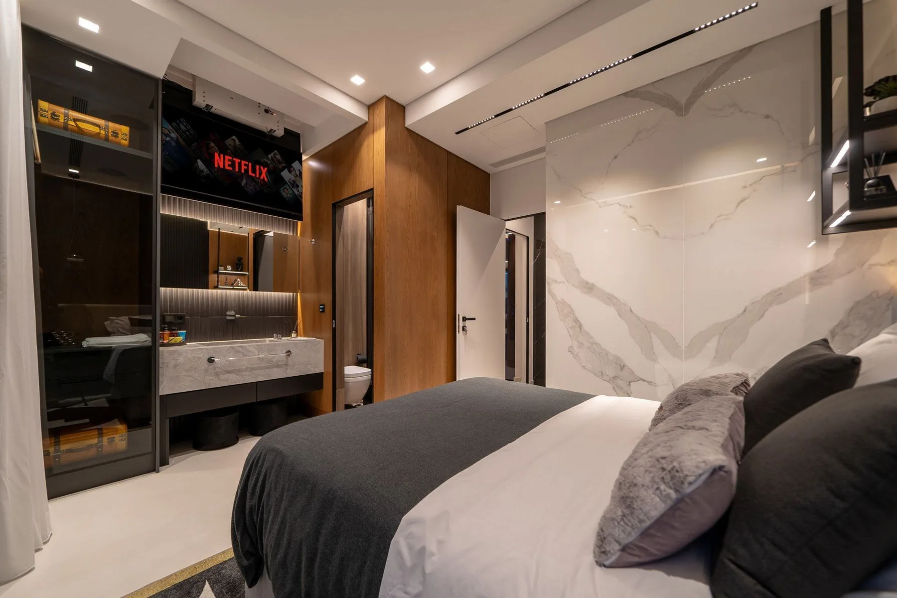 A modern bedroom features a dark upholstered bed, a marble-patterned wall, and a built-in vanity with a television.