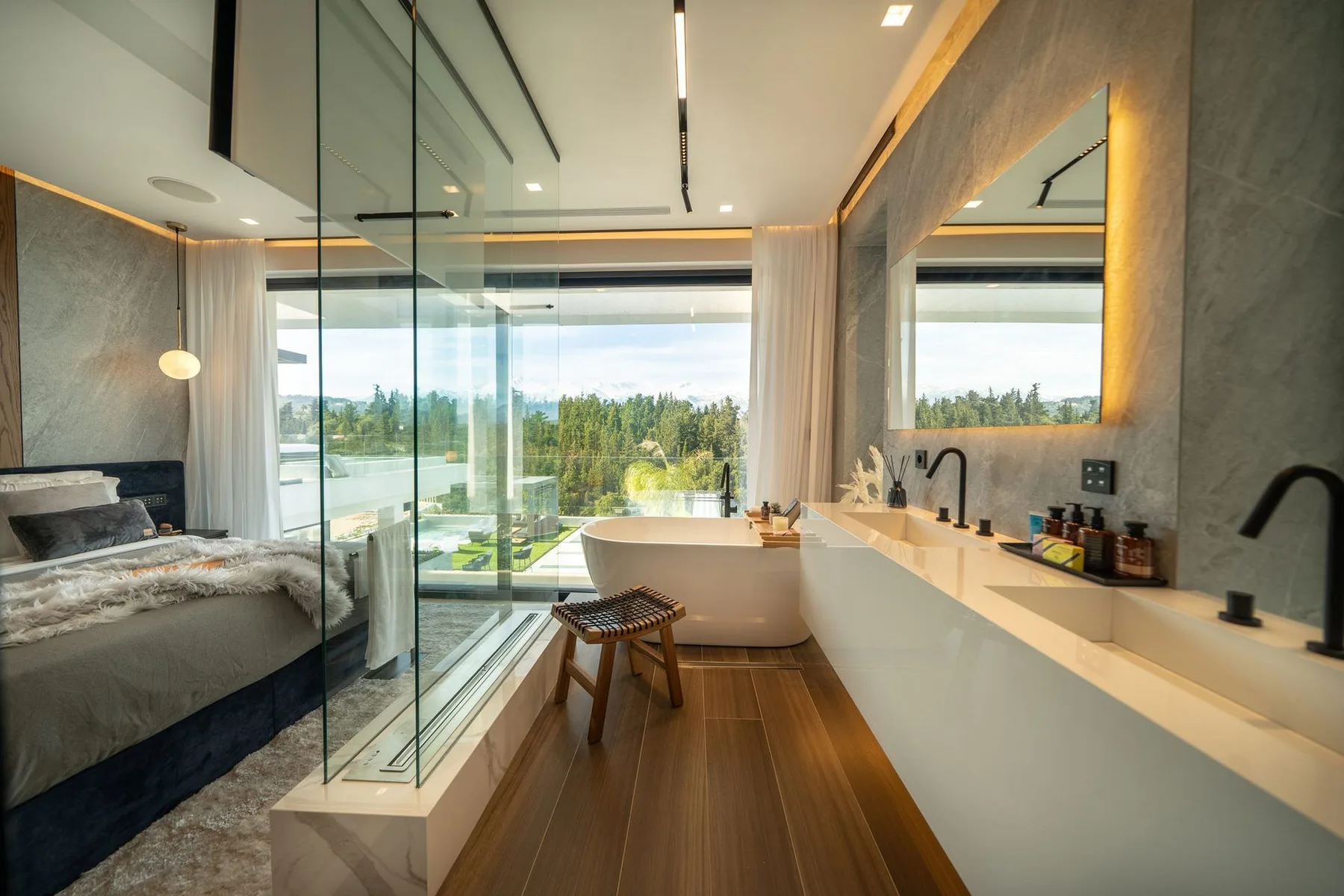 A modern bedroom and bathroom share a glass partition, with a freestanding tub, long vanity, and large windows.