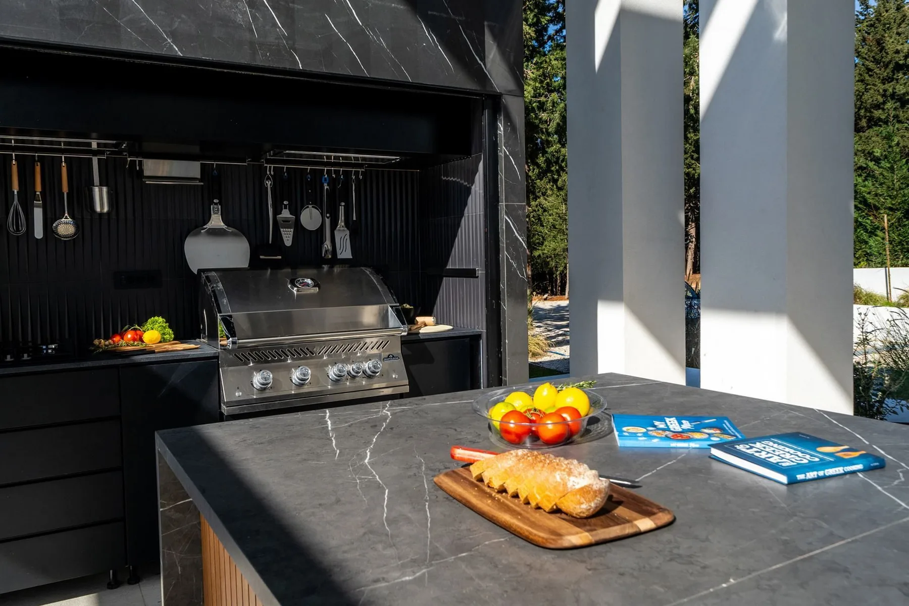An outdoor kitchen features a stainless-steel grill, hanging utensils, and a dark stone island with fruit and bread.