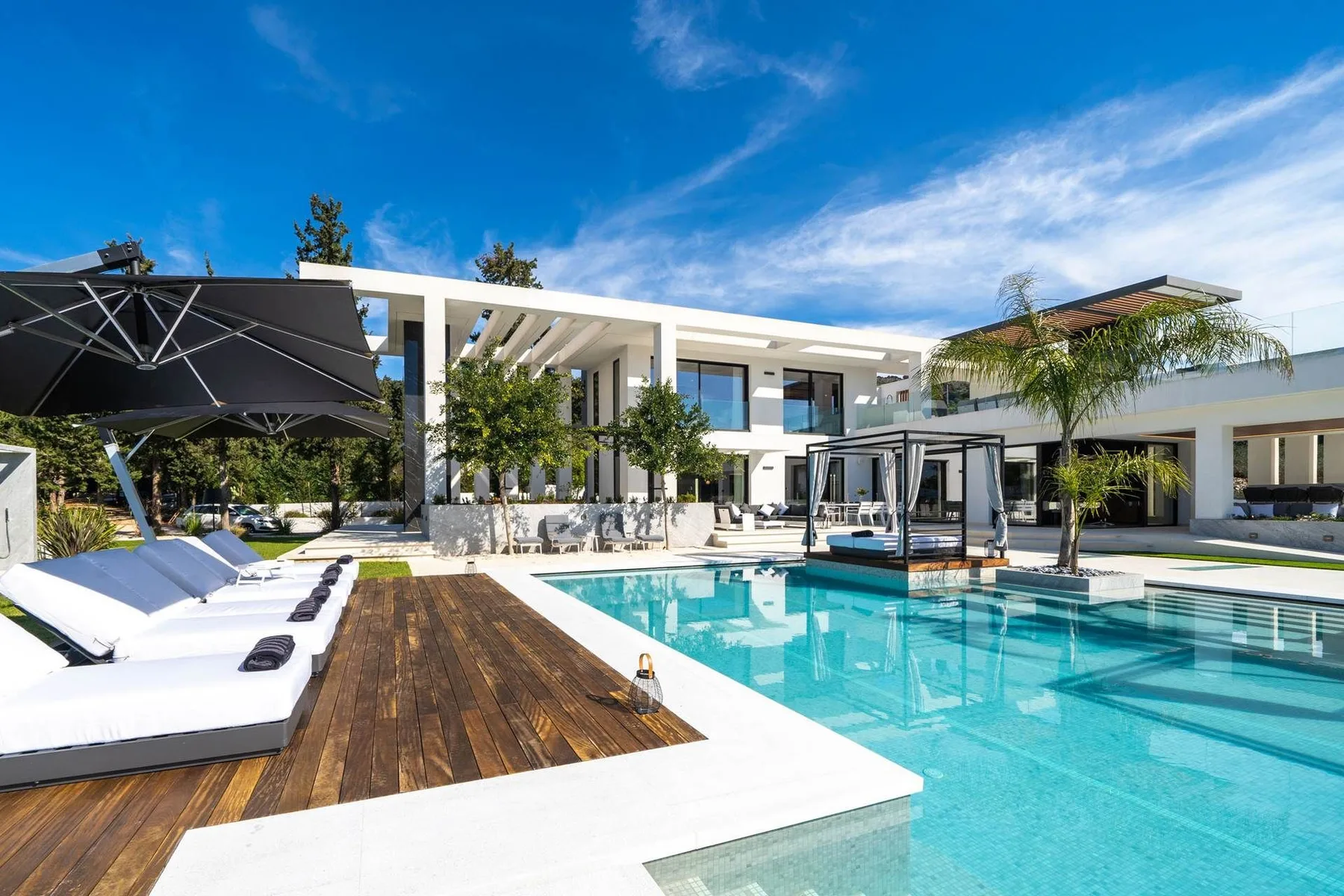 A modern white villa surrounds a turquoise swimming pool, with wooden decking and white loungers.