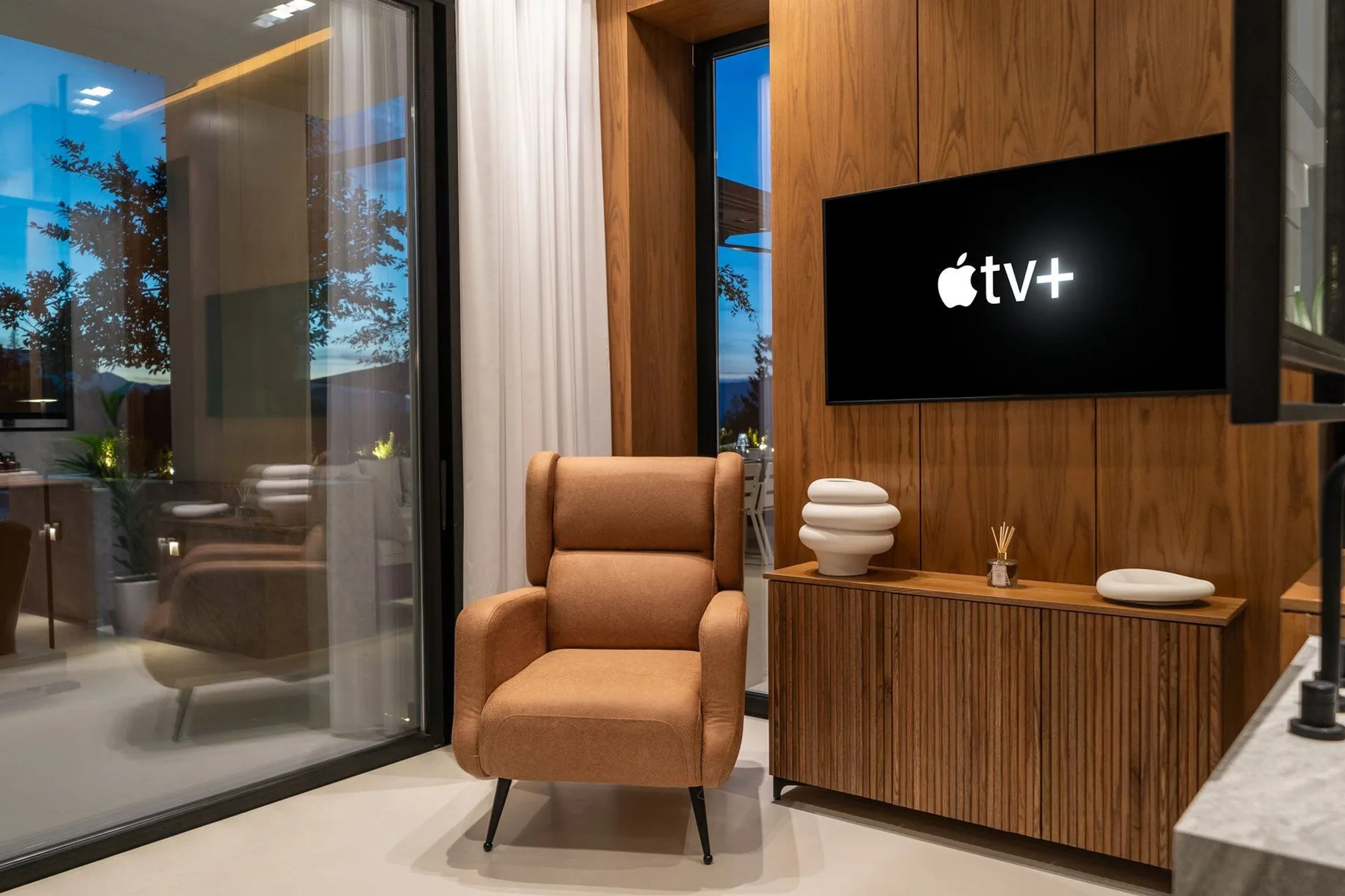 A tan armchair sits beside a wood-paneled television console, with floor-to-ceiling glass doors nearby.