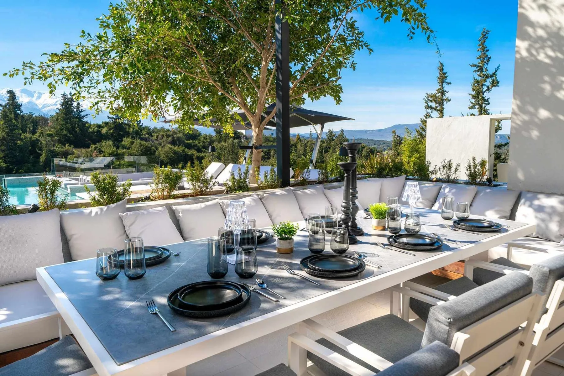 An outdoor dining table is set with dark plates beside cushioned seating, a swimming pool, and leafy trees.