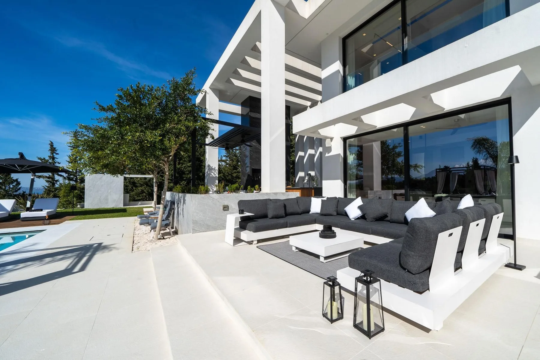 A modern white villa opens onto a patio with dark outdoor sofas, a dining table, and a swimming pool.