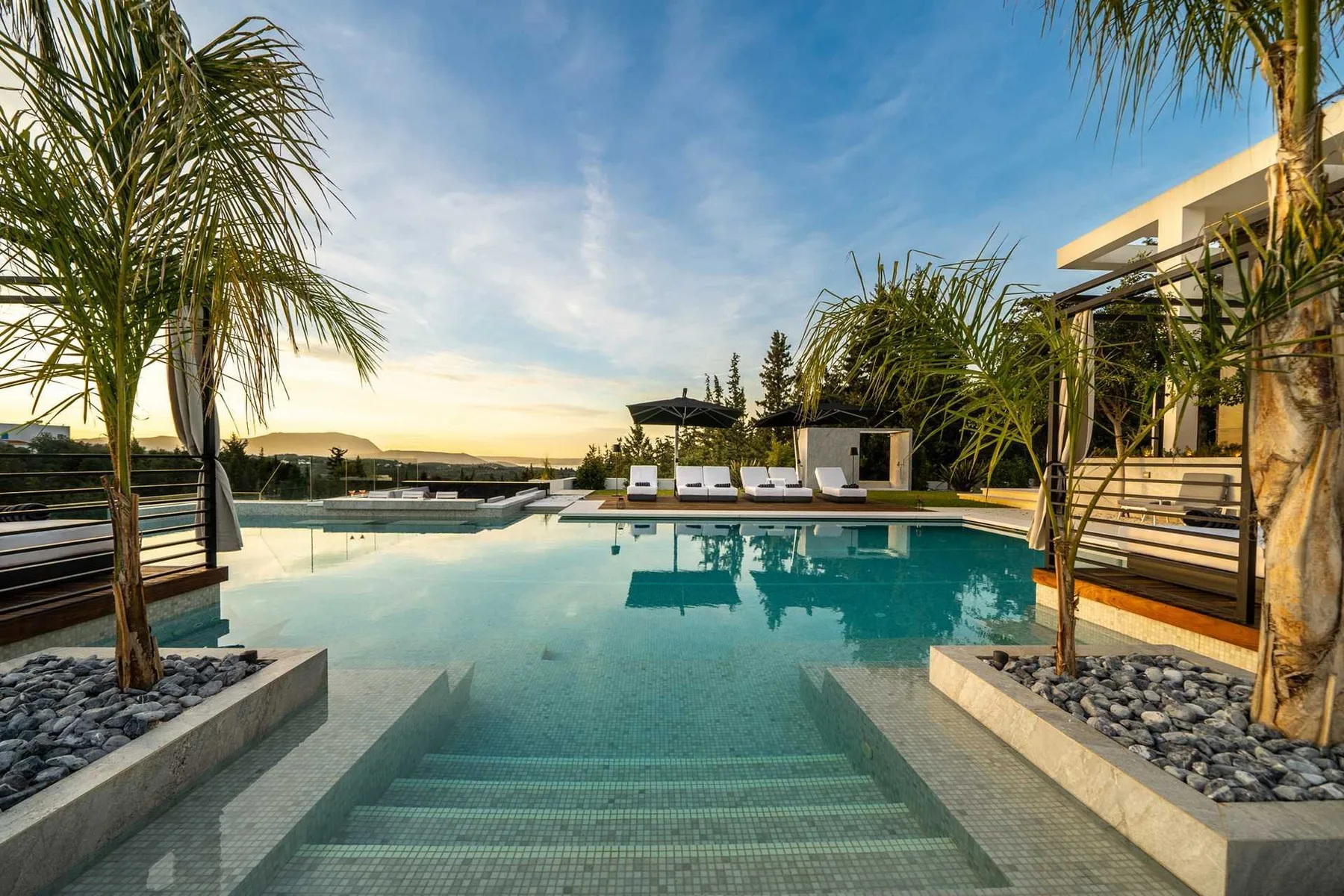 A turquoise outdoor pool has broad submerged steps, lounge chairs, and palm trees around its edges.