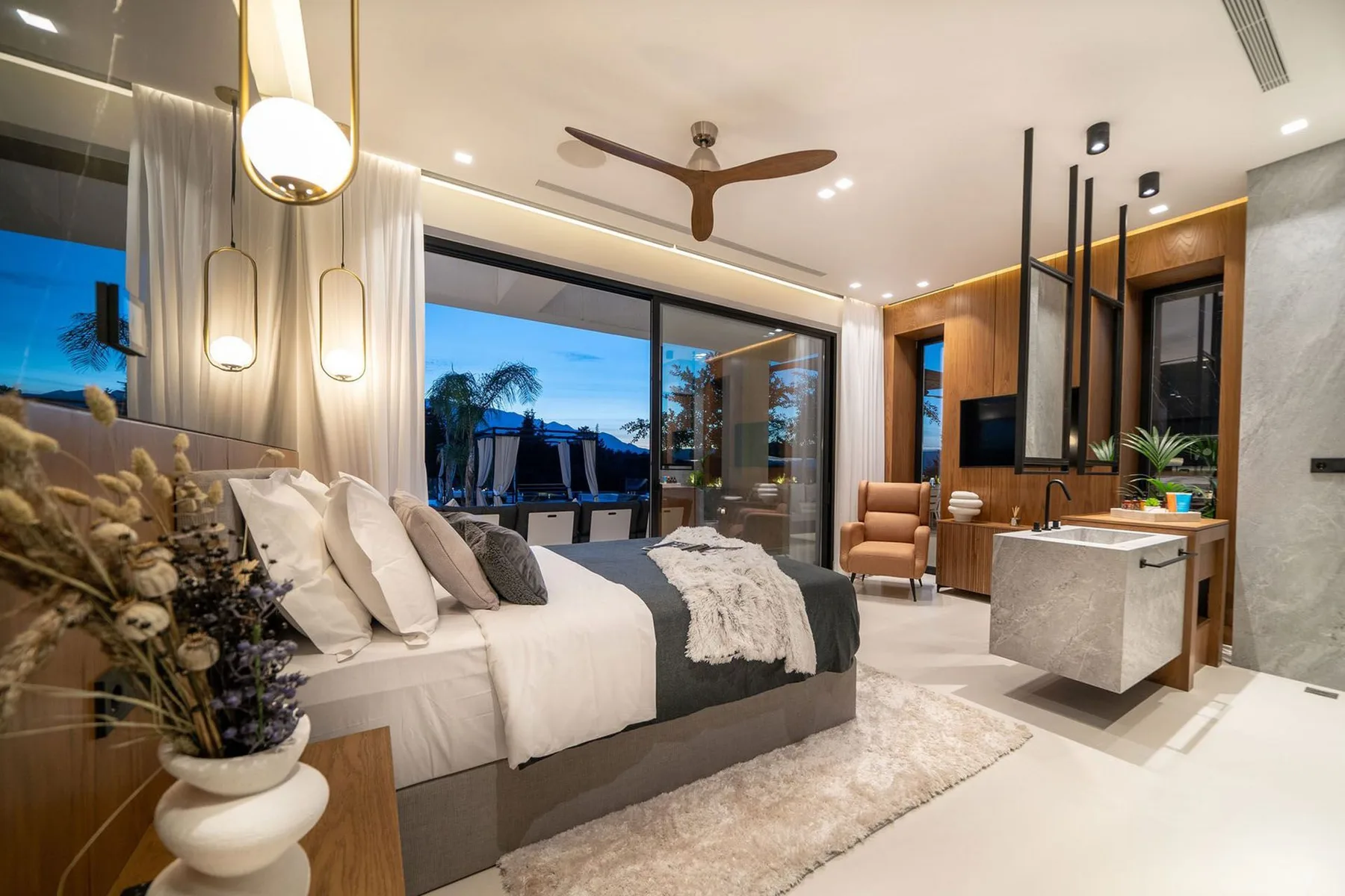 A modern bedroom features a king bed, floor-to-ceiling glass doors, and a stone-topped vanity.