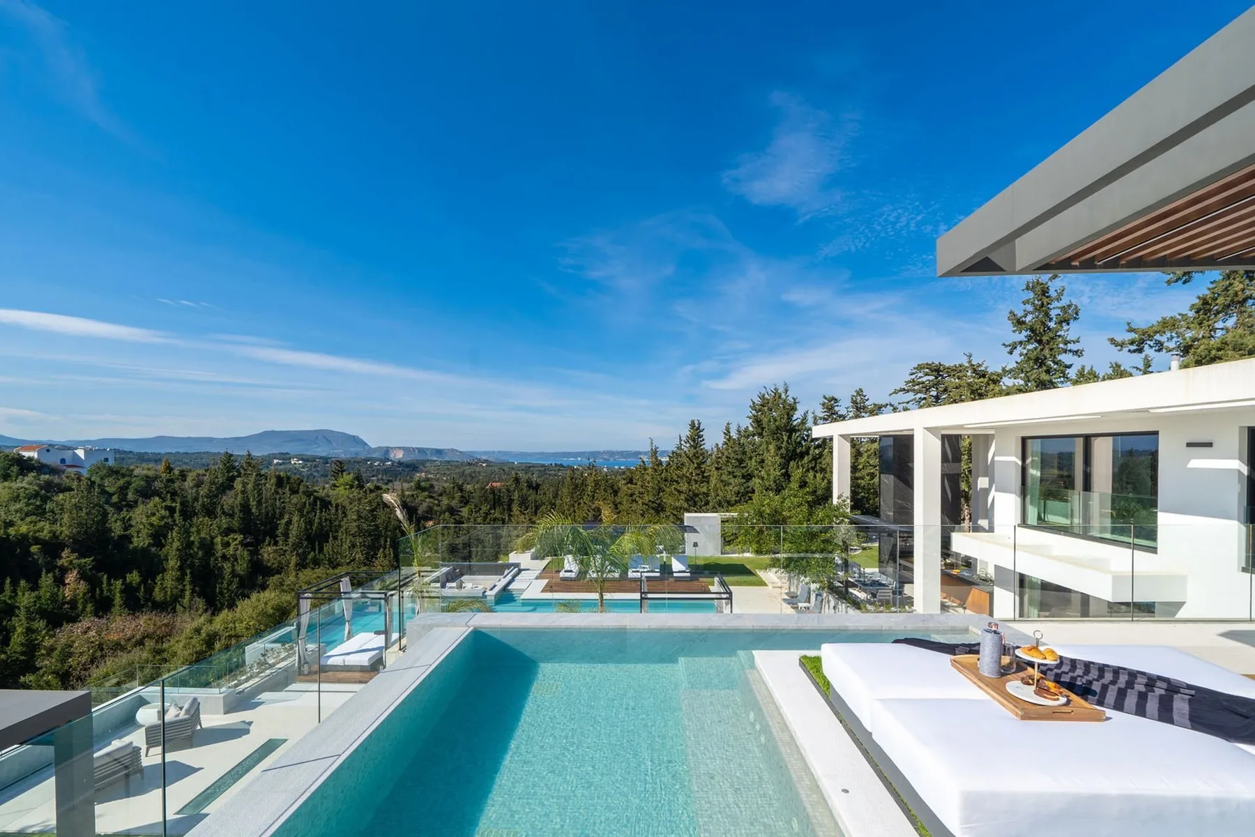A modern hillside house overlooks a turquoise swimming pool, glass railings, and a wooded landscape.