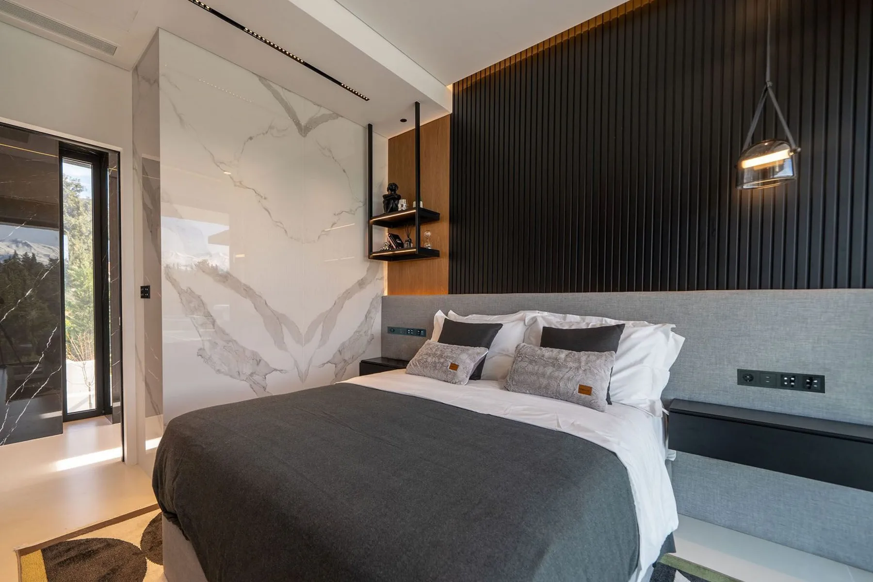 A modern bedroom features a large bed with gray bedding, a marble-patterned wall, and dark vertical paneling.