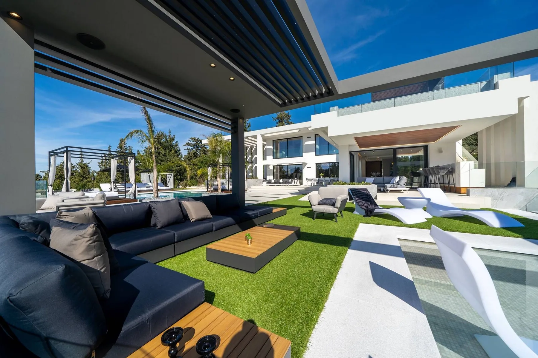 A modern white villa opens onto a covered patio with dark sofas, a green lawn, and sculptural loungers.