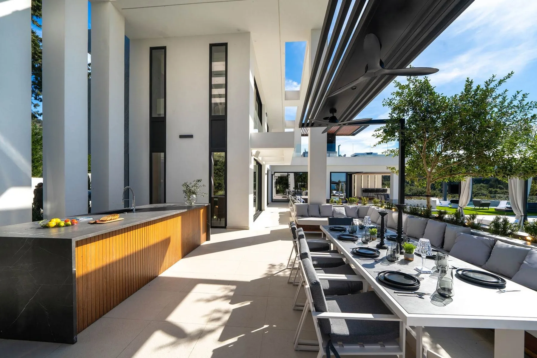 A modern outdoor dining area features a long table, cushioned chairs, and a shaded counter beside the garden.