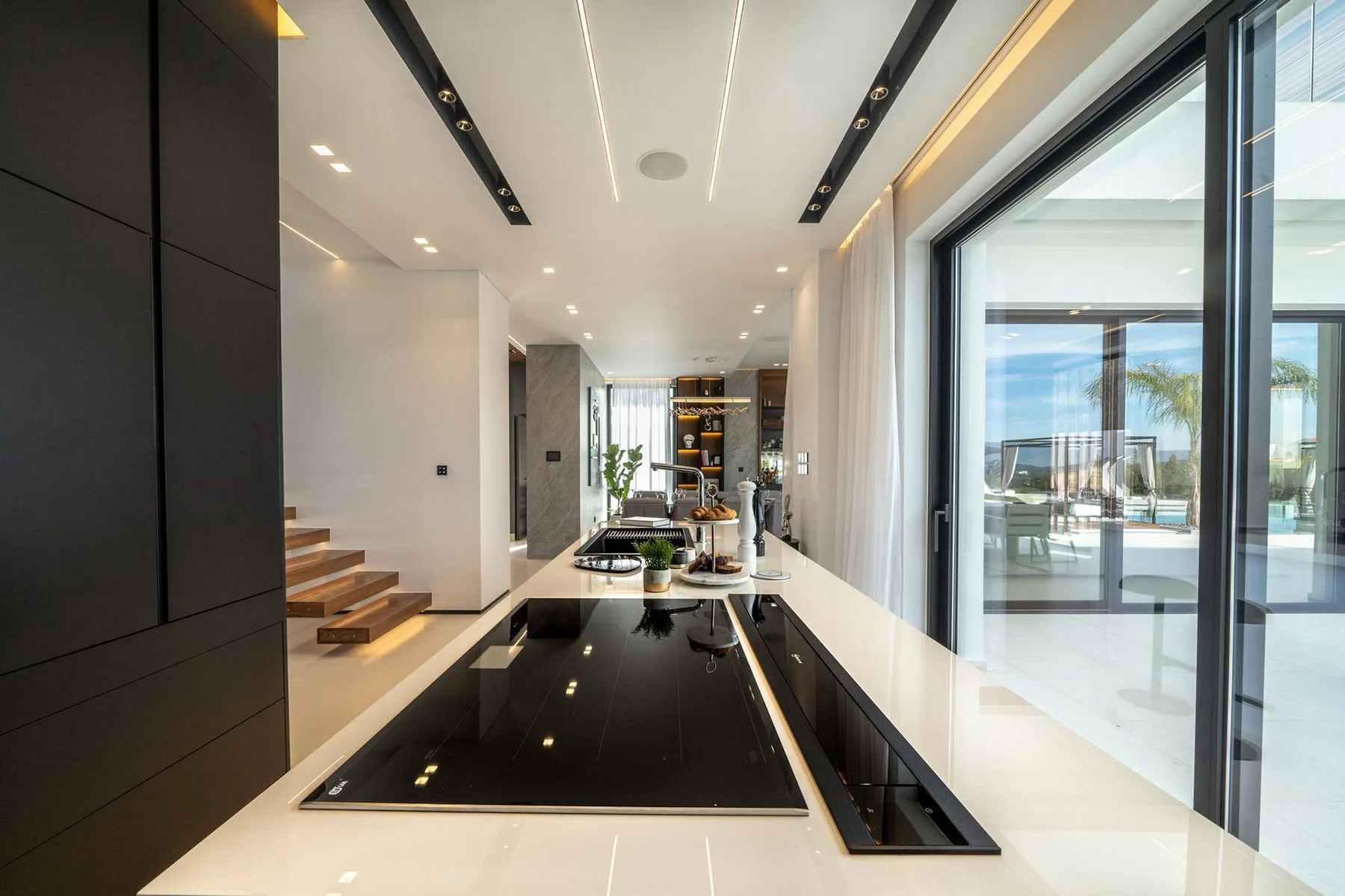 A modern kitchen with a long black island, wood steps, and floor-to-ceiling glass doors.