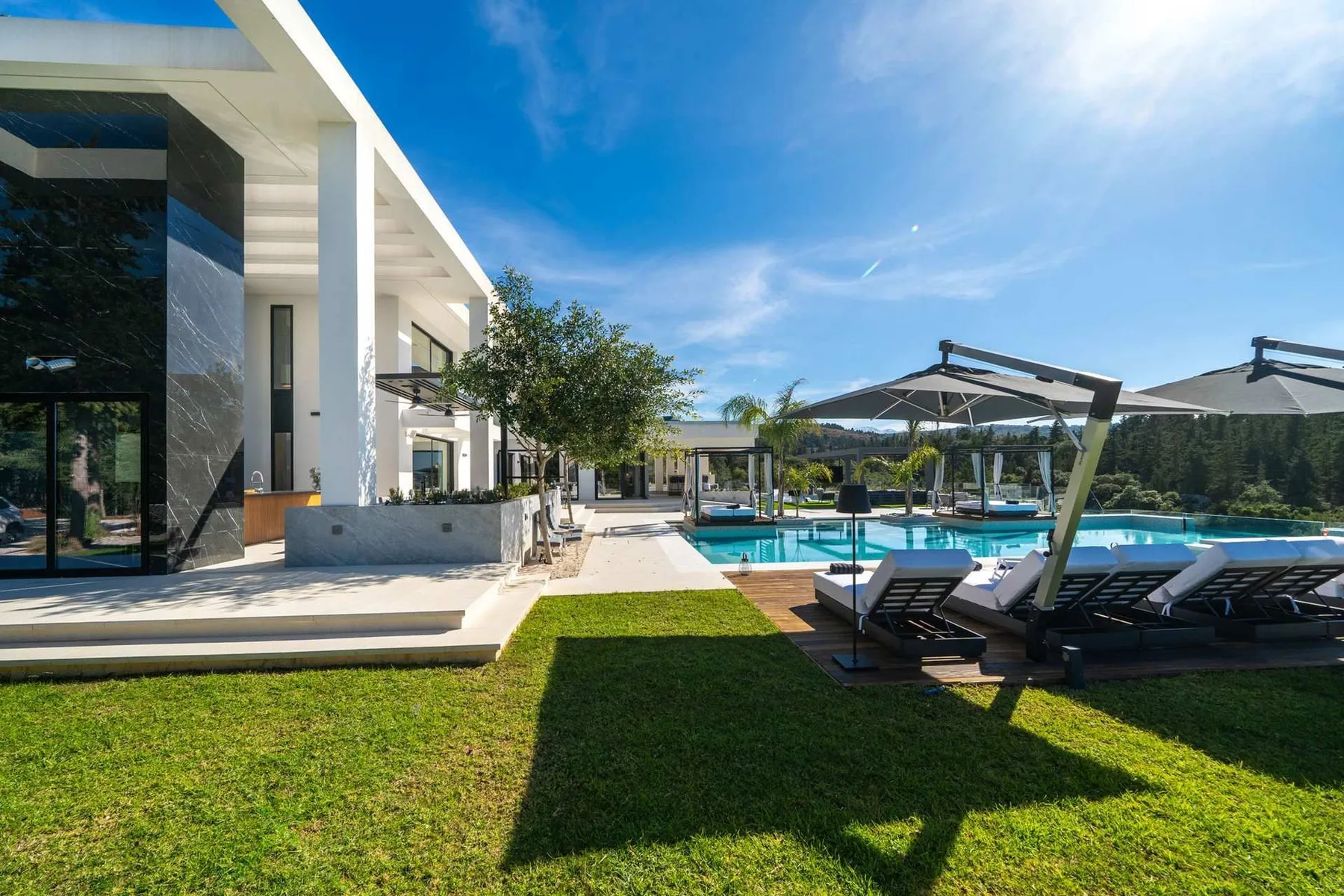 A modern white building overlooks a swimming pool, with sun loungers and umbrellas beside a green lawn.