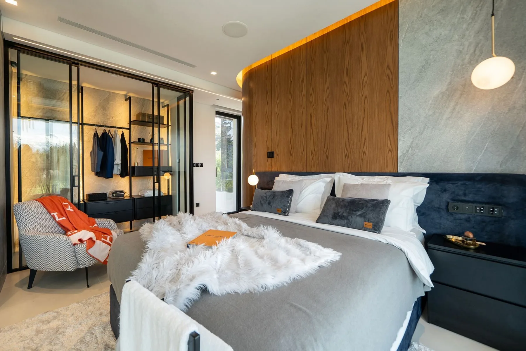 A modern bedroom with a gray bed, white pillows, and a glass-fronted wardrobe.
