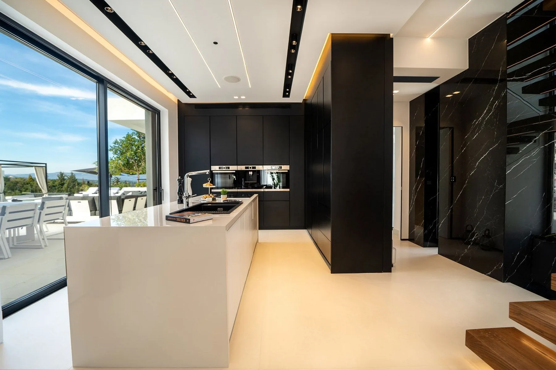A modern kitchen features dark cabinetry, a pale island, and large windows opening onto a terrace.