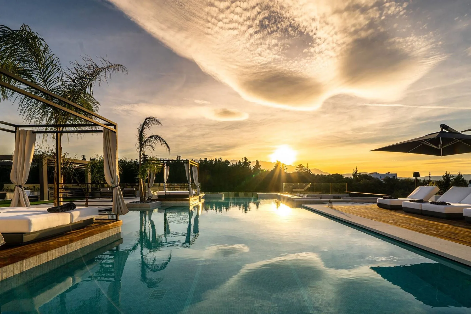 A swimming pool reflects the sunset beside lounge chairs, shaded cabanas, and palm trees.