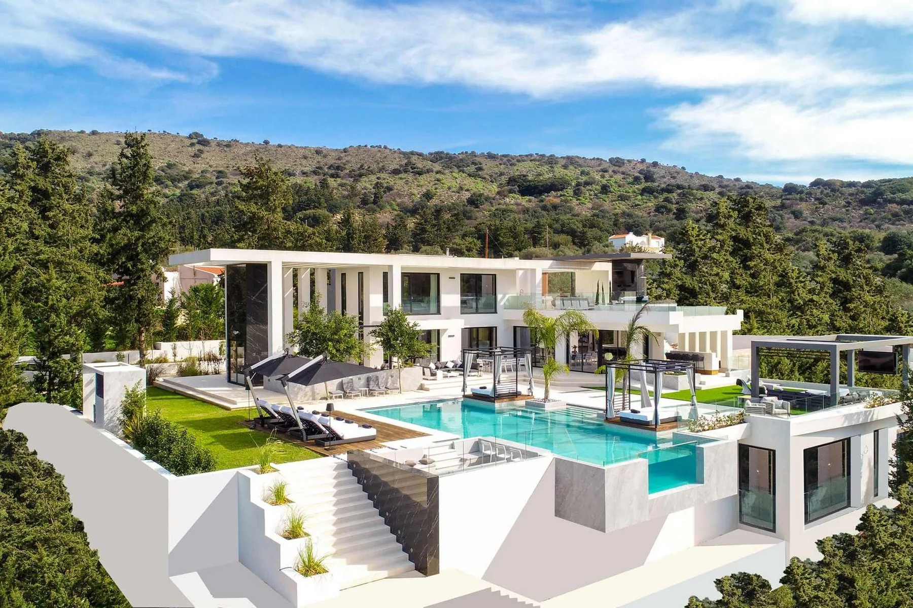 A modern white villa overlooks a turquoise swimming pool, with lounge chairs, terraces, and wooded hills beyond.